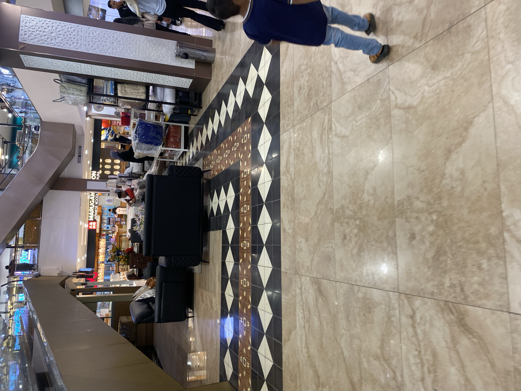 The image shows an interior scene within a shopping mall in Banda Aceh, Indonesia. The foreground is dominated by large, beige marble tiles. A decorative band of smaller tiles, featuring a diamond pattern in black, white, and brown, runs horizontally across the mid-ground. In the background, a young boy wearing a dark blue t-shirt and blue jeans walks away from the camera, his feet clad in dark shoes. To the left of the boy, there are two dark-colored sofas. Further in the background, a store display features shelves stacked with bedding items like duvets and pillows. Beyond that, there is a store with the sign "MARUGAMI JAPANESE NOODLE" visible. People are seen walking and browsing in the mall. An escalator is visible on the left, leading to an upper level. The overall environment is that of a modern retail space.