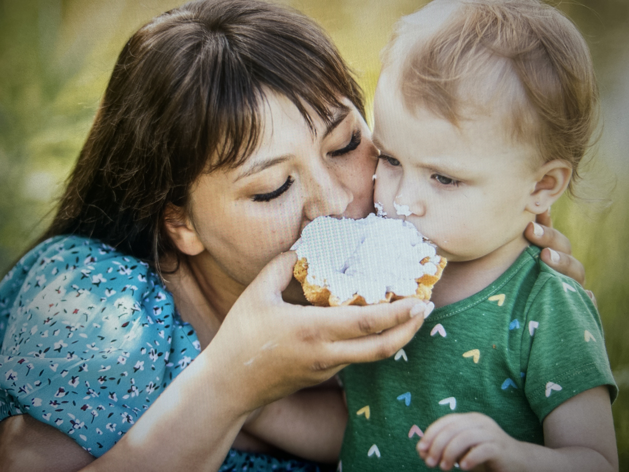 This media file shows a close-up, softly-lit outdoor portrait of a woman and a young child. The woman, with dark hair and closed eyes, is leaning in to kiss or share a treat with the child. She is wearing a blue dress with a small floral pattern. The child, who appears to be a toddler with blonde hair, is looking downwards with a somewhat serious expression. They are wearing a green t-shirt adorned with a pattern of colorful heart shapes. The focus is on the interaction between the two, with a blurred background of green foliage suggesting a natural, outdoor setting, possibly a park or garden. The lighting and soft focus create a warm and intimate atmosphere, hinting at a moment of affection and connection. The treat being shared appears to be a small, round pastry with white frosting. The scene is likely set during the daytime, given the natural light. There is no visible text in the image.