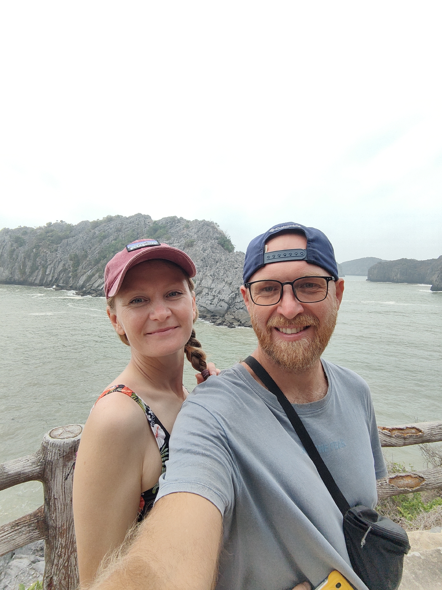 A selfie captures a man and a woman on a lookout point in Hải Phòng, Vietnam. The woman on the left, is light-skinned with blonde hair in a braid, wearing a dark-floral-print top and a pink baseball cap with a visible logo. The man on the right is fair-skinned with red hair and a beard, wearing a light-blue T-shirt, black-framed glasses, and a navy baseball cap; he has a black crossbody bag. Behind them, the scene consists of choppy water and limestone islands under an overcast sky. A rustic wooden railing is visible at the edge of the lookout point.