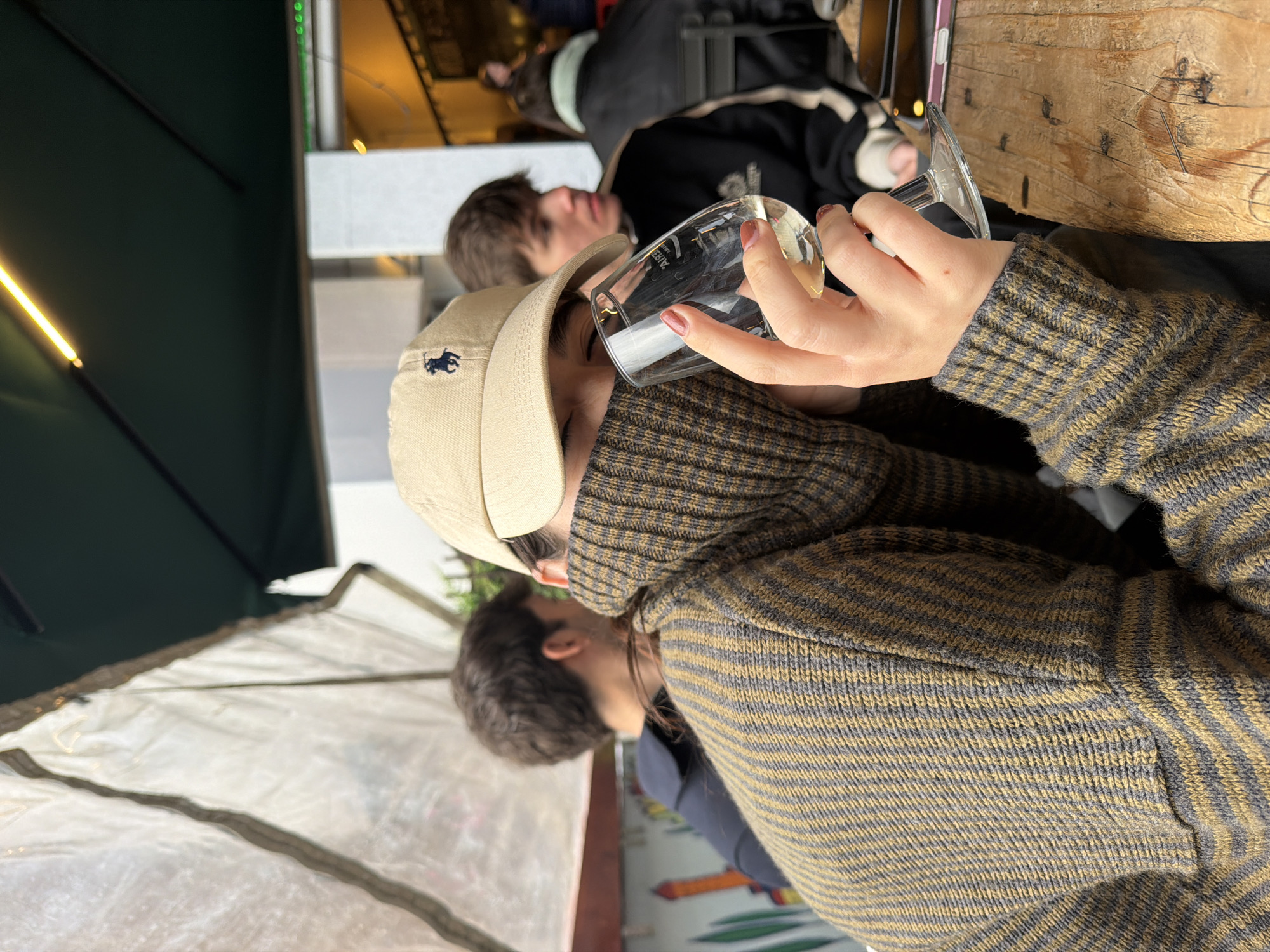 A medium shot shows a person in a beige cap and brown striped sweater holding a wine glass with their right hand up to their face, likely drinking, while sitting at an outdoor table in Milan, Italy. Another person is behind them, and the table is made of wood. A clear tent is to the side, and a green awning is above.