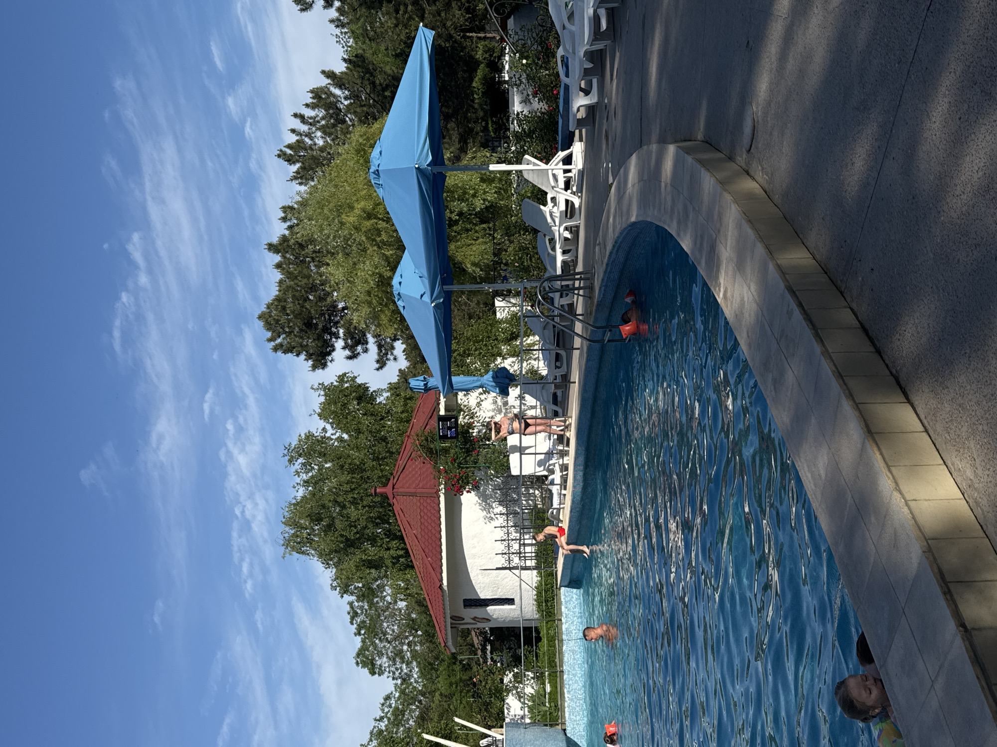 The image shows a swimming pool scene, presumably during daytime, in what appears to be Yevpatoriya, Ukraine. The pool is surrounded by a tiled deck with lounge chairs and blue umbrellas. Several individuals are present in and around the pool; some are swimming while others are standing near the edge. There is a small white building with a red roof near the pool area, partially obscured by trees. The sky is blue with scattered clouds.