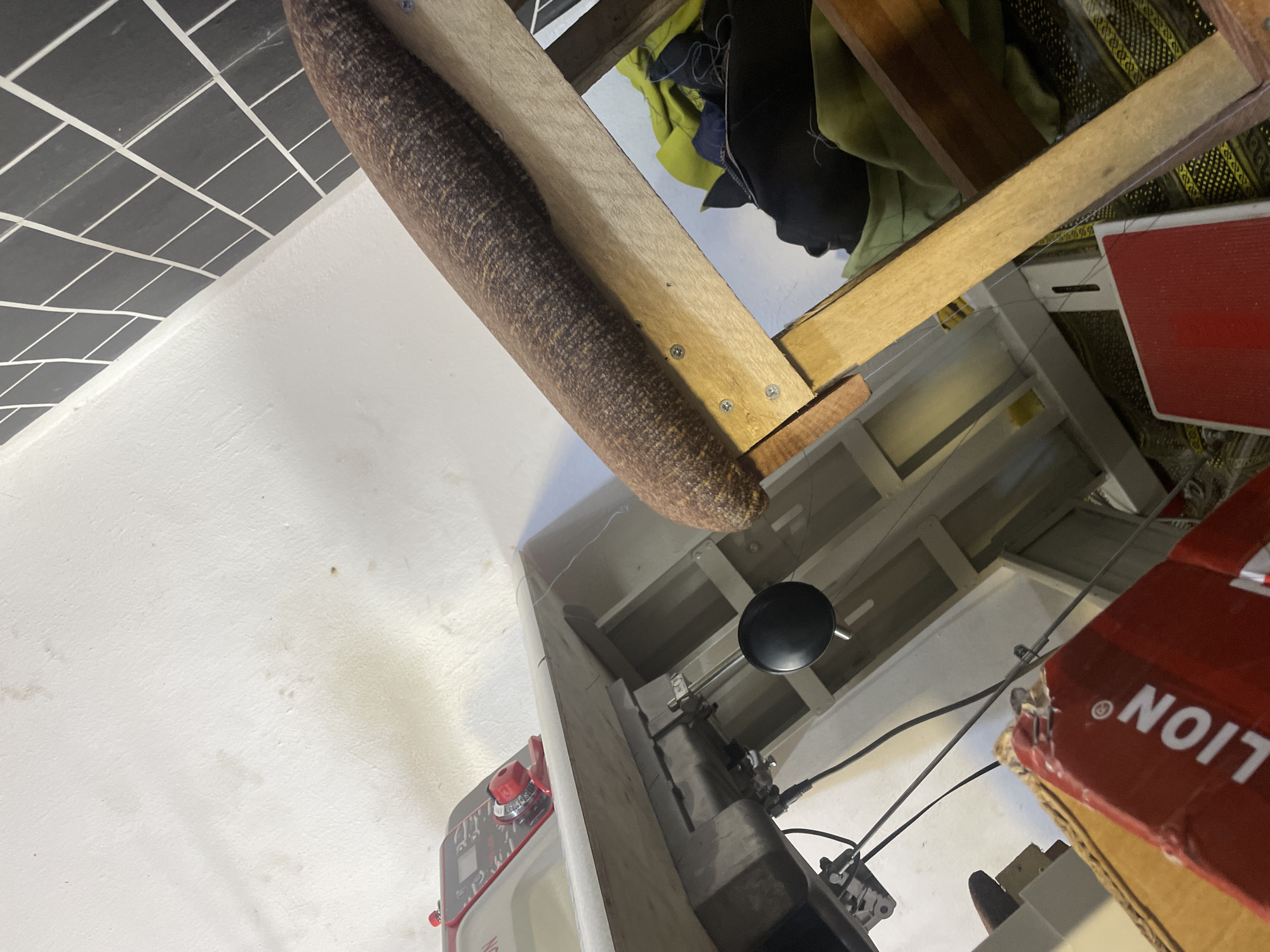 The image shows an indoor scene, possibly a workshop or storage area, with a focus on various objects. In the foreground, a white wall is visible with some light staining. To the left, part of a red and gray appliance with a digital display and a red knob is seen. A brown, cylindrical object with a textured surface hangs from a wooden structure above. This wooden structure, made of planks, is supported by what appears to be a shelf system. Behind the wooden structure, several pieces of fabric in green and black are draped.
Below the wooden structure, a black spherical object, possibly a light fixture or part of a machine, hangs from a wire. A sewing machine or a similar mechanical device is partially visible, with black wires and metal components. To the right, a red box with white text is prominently featured. The visible text includes a circular symbol and the word "LION" printed backward. The overall impression is one of a cluttered but functional space. The lighting suggests it is daytime, but no specific time or weather conditions can be determined. No people are visible in the image. The location context provided is Jalingo, Nigeria.