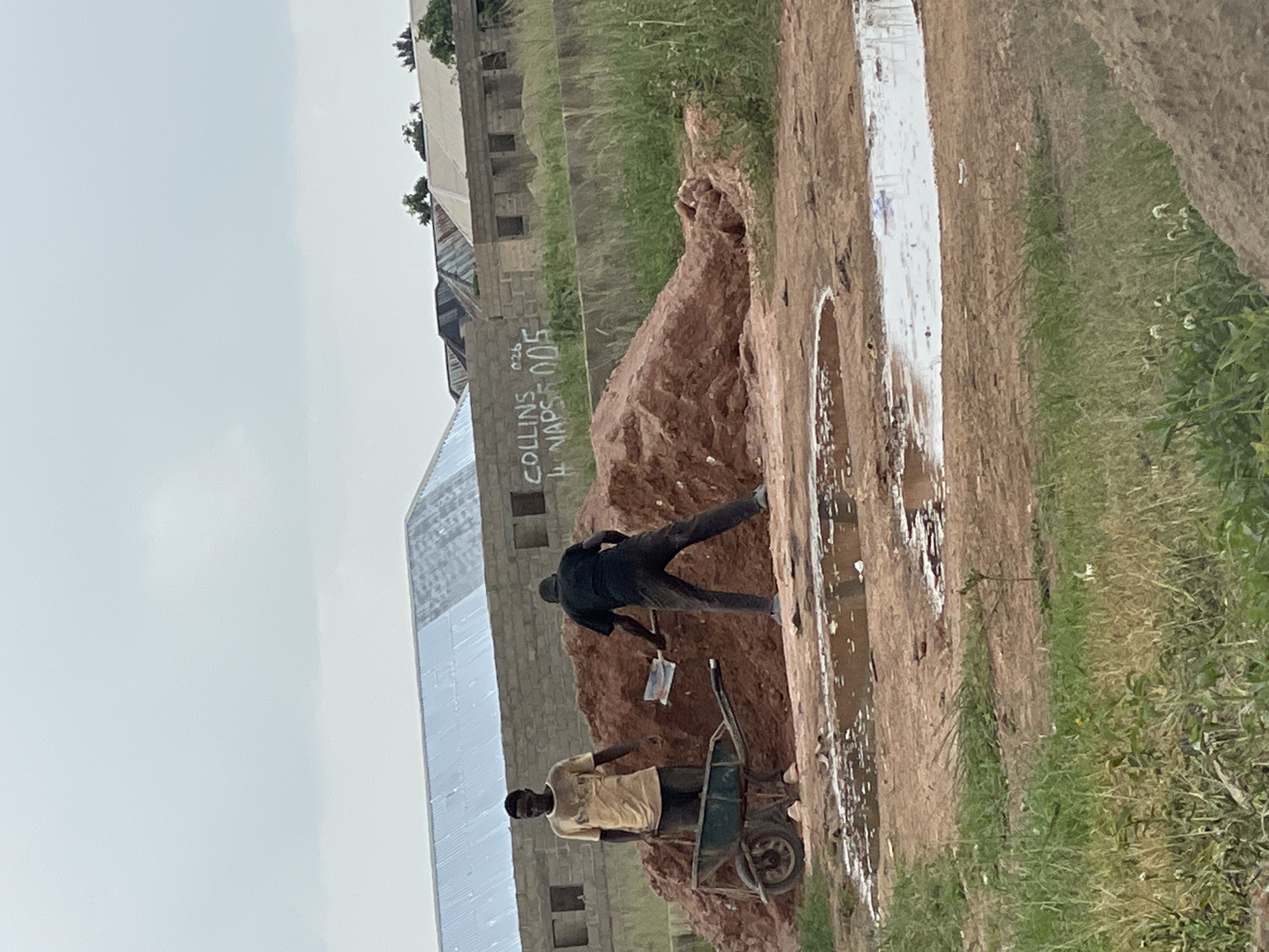 Two men are working at a construction site in Durumi, Nigeria, a suburb of Abuja. The man on the left, wearing a yellow t-shirt and dark pants, stands beside a wheelbarrow. He is looking at the other man. The man on the right, wearing a black t-shirt and dark pants, is using a shovel to dig into a large mound of red dirt. The background shows partially constructed buildings with corrugated metal roofs. The ground in the foreground is muddy and has several puddles of water, suggesting recent rain. There are signs spray-painted on a wall in the background that read "COLLINS 026" and "4 NAPS 005".