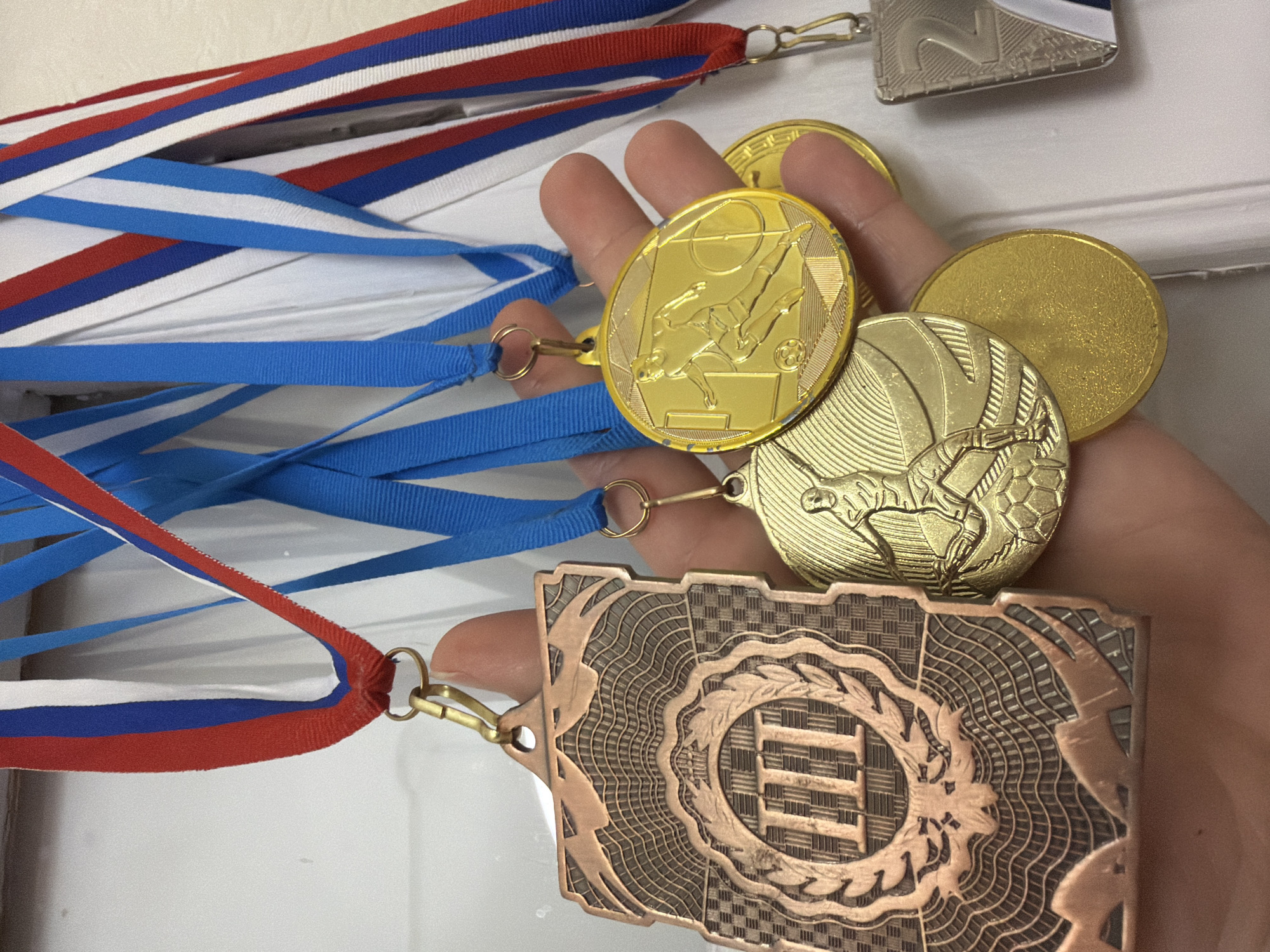 A hand holds several medals, including a bronze medal, two gold medals with a soccer player and horseback rider on them, and a silver medal. The medals have red, white, and blue ribbons. The location is not discernible from the image.
