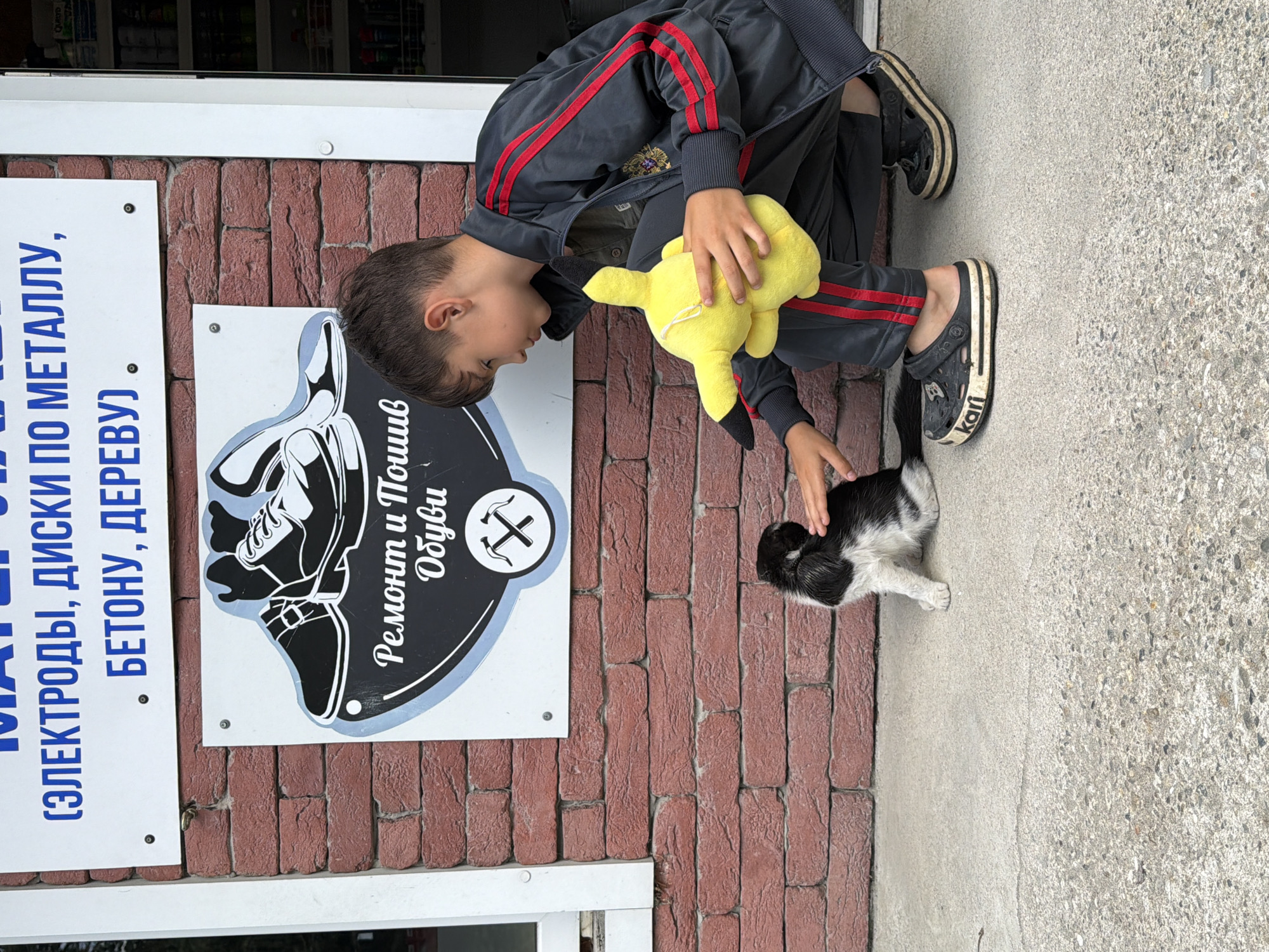 A young boy is crouching on a concrete sidewalk in front of a brick building. He is wearing a dark grey tracksuit with red stripes down the sleeves and legs, and black clogs. In his left hand, he is holding a yellow plush toy. His right hand is extended and touching a small black and white kitten, which is sitting on the sidewalk. Behind the boy and kitten is a sign with a picture of shoes and the text "Ремонт и Пошив Обуви" (Shoe Repair and Sewing). Above this sign, another sign reads "(ЭЛЕКТРОДЫ, ДИСКИ ПО МЕТАЛЛУ, БЕТОНУ, ДЕРЕВУ)" (Electrodes, metal, concrete, wood discs). The building appears to be a shop or service establishment in the city of Shepsi, Russia.