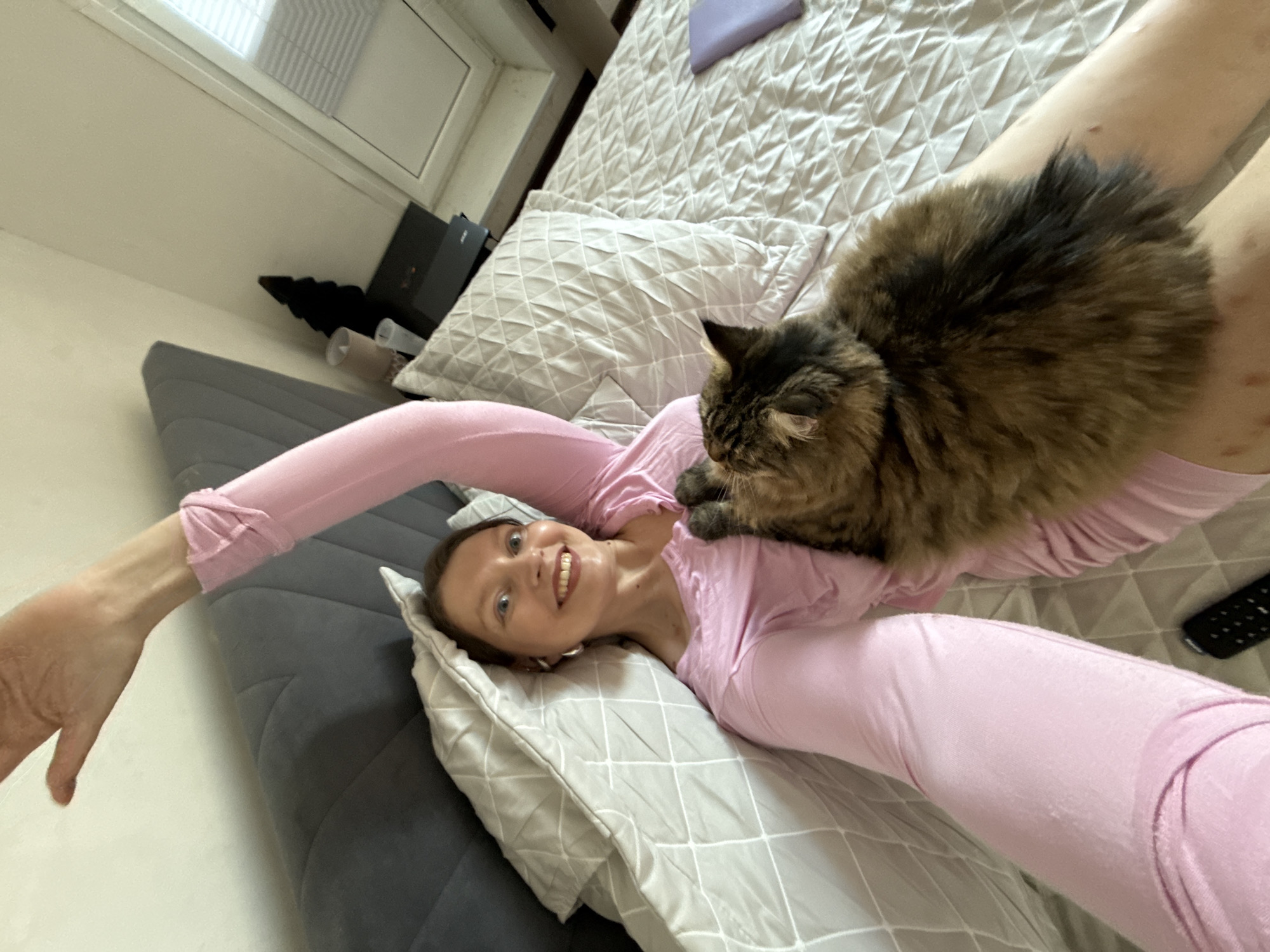 A selfie taken indoors shows a woman lying on her back in a bed with a cat on top of her. The woman has fair skin, light eyes, and is smiling with visible teeth. She is wearing a pink long-sleeved shirt. A fluffy, medium-sized, brown and black tabby cat is resting on her chest. The bed has a gray headboard and a white, quilted comforter. There are multiple pillows on the bed, also quilted. To the left of the bed is a window and a black object on a shelf. A remote control is visible in the bottom right corner. The location is not discernible.