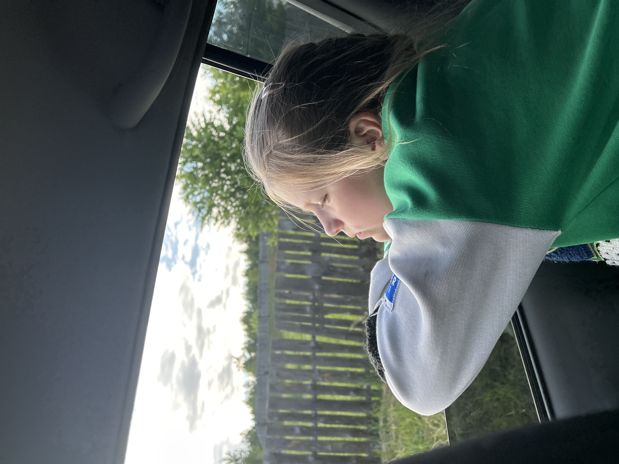 A young blonde girl is looking out the window of a vehicle. She is leaning her head on her arms, which are crossed in front of her. Her hair is light blonde and appears to be blowing in the wind. She is wearing a green and white sweatshirt. The background shows a wooden fence, trees, and a cloudy sky. The image is taken from inside the vehicle, looking out. The location of this scene is the city of Fokino, Russia, though this detail is not visually apparent in the image itself.