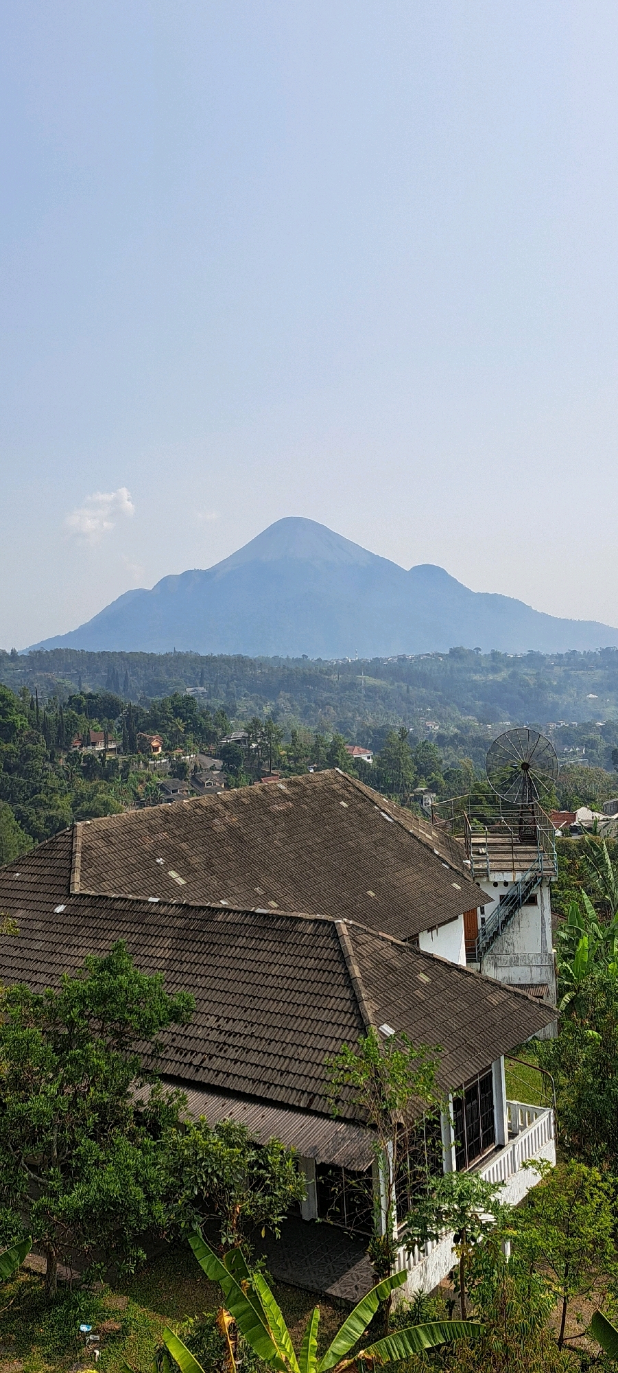 The image captures a panoramic view from an elevated position overlooking a residential area with a prominent mountain range in the background. In the foreground, two distinct tiled rooftops of houses are visible. The larger, closer roof is a dark brown, textured material. To its right, a smaller, white structure with a similar dark tiled roof is partially visible. Adjacent to the white structure is a tall, white tower with an industrial satellite dish mounted on top, accessed by a metal staircase. The middle ground is a lush, green landscape dotted with various houses, trees, and winding roads, suggesting a suburban or semi-rural environment. The background is dominated by a hazy, blue mountain range, with a distinct, large peak at the center. The sky is a clear, pale blue with a faint wisp of a cloud. The scene appears to be in Prigen, Indonesia, as indicated by the context. There are no people visible in the image. The environment is characterized by dense vegetation, residential structures, and natural topography.
