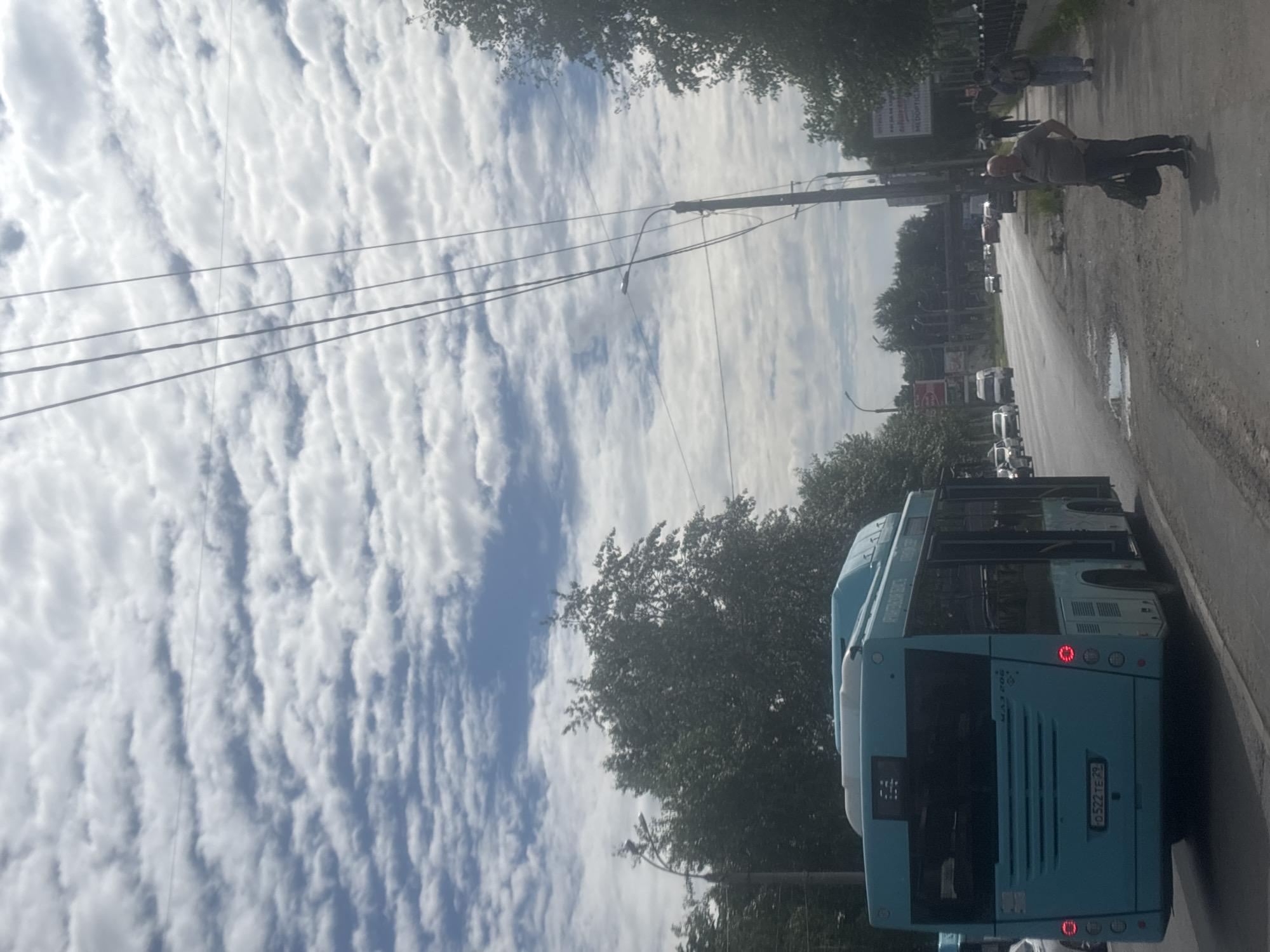 The image captures a street scene in Arkhangelsk, Russia, under a sky filled with a textured pattern of white clouds. A turquoise bus, identified by the license plate "0522TE 29," is prominently positioned on the left side of the frame, facing away from the viewer and traveling down a paved road. To the right of the road, a man stands on a sidewalk, holding a dark bag in his left hand and with his right hand on his hip. Another person, partially obscured by the man, walks away from the viewer on the same sidewalk. The road is lined with trees and utility poles, and a sign is visible on a pole in the distance. The overall environment suggests an urban setting on a cloudy day.