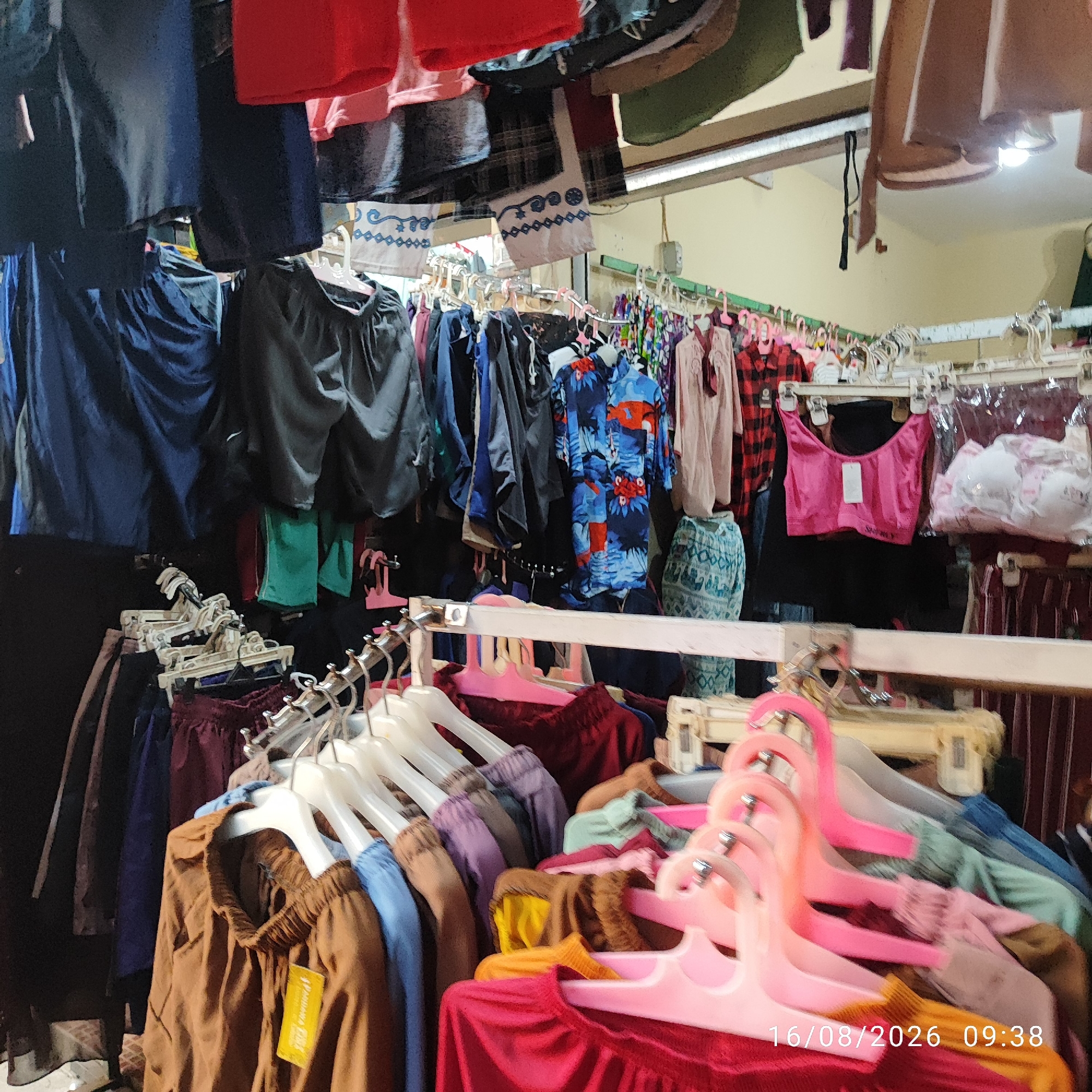 The image depicts a clothing store in a densely packed market setting. Various items of clothing, including shorts, shirts, and what appear to be undergarments, are displayed on racks and hangers. The clothing is varied in color and pattern, with some items exhibiting a more casual style and others appearing more formal. The environment seems to be indoors, with artificial lighting casting shadows on the merchandise. The overall impression is of a bustling marketplace where a wide array of textiles are being offered for sale. The scene is static, with no people visible in the frame. The date stamp at the bottom of the image indicates "16/08/2026 09:38".