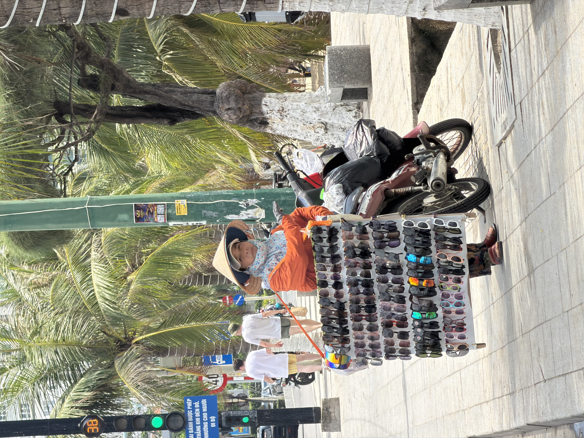 This image captures a bright, sunlit street scene in Nha Trang, Vietnam, featuring a street vendor and passersby. In the foreground, a person, likely a woman, wears a traditional Vietnamese conical hat (nón lá) and an orange top over a patterned shirt. She sits behind a large display unit covered with numerous pairs of sunglasses of various designs and colors, offered for sale. Behind her, a motorbike is parked, heavily loaded with black plastic bags and other items.
Two men, dressed in white t-shirts and shorts, are seen walking along the paved sidewalk, passing by the vendor's stall. The background is dominated by lush green palm trees, indicative of a tropical climate. To the left, a green traffic light displays the number "38," and a blue road sign is visible on a pole. The text on the blue sign, in Vietnamese, reads: "XE ĐẠP ĐƯỢC PHÉP THẲNG KHI ĐÈN ĐỎ. NHƯỜNG CHO NGƯỜI ĐI BỘ," translating to "BICYCLES ARE ALLOWED TO GO STRAIGHT ON RED LIGHT. YIELD TO PEDESTRIANS." Other smaller advertising posters with partial text like "SE" are also affixed to a green pole. The scene reflects a typical daytime street market or vending activity in a bustling urban environment.
