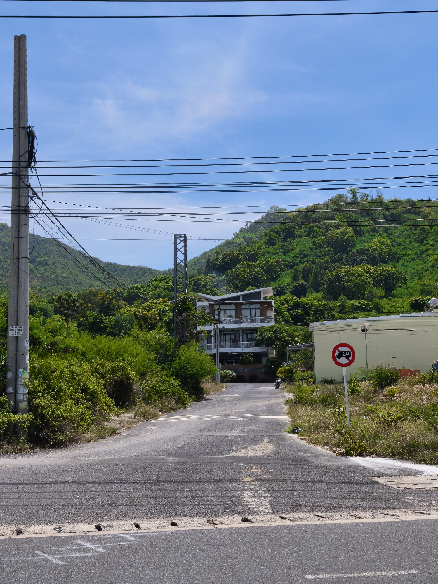 The image depicts a road in Bac Nha Trang Ward, Vietnam, leading towards a lush green hillside. The road is paved and shows signs of wear. On the left side of the road, a tall utility pole with numerous wires is prominent. Further down the road, a modern multi-story building with balconies is visible, surrounded by dense green vegetation. To the right of the road, a yellow building is present, and a circular red and white sign indicates a weight limit of 2.5 tons for vehicles. A pedestrian crossing is marked on the road ahead. The sky is bright blue with scattered clouds, and the overall environment suggests a sunny day.