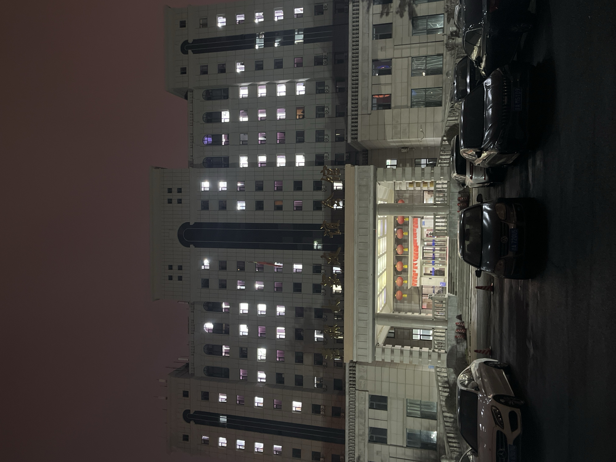 The image shows a tall, light-colored building at night, likely located in Changchun City, China. The building is illuminated, with many windows lit. Several cars are parked on the street in front of the building. The sky is a dark, reddish-gray, suggesting twilight or a polluted atmosphere. There are few people visible, and the scene appears quiet. The architecture of the building suggests it could be a government office or a commercial building. Decorations adorn the entrance, implying it is an important location. Traffic cones are set up near the entrance. The photo is angled, giving a slightly distorted perspective. The image does not offer enough information to confirm a specific location.