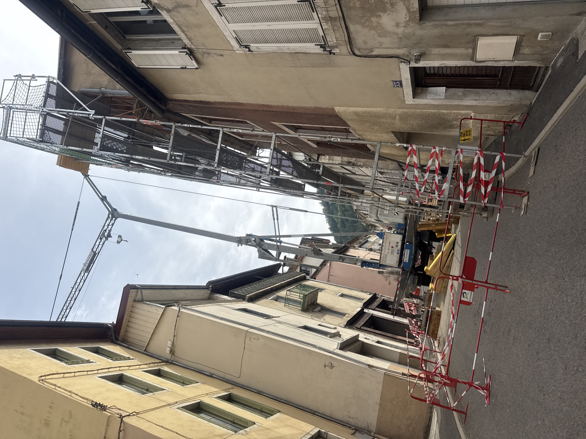 The image depicts a narrow street in the city of Saint-Claude, France, lined with buildings that appear to be under renovation or construction. A large metal scaffolding structure is erected along the side of one building, reaching several stories high. A crane, possibly part of the construction equipment, is visible, extending over the street. Red and white striped temporary barriers are set up on the street, indicating a closed-off or restricted area. The street is paved and appears to be relatively quiet, with no visible people or vehicles other than the construction equipment. The sky is overcast, suggesting a cloudy day. The architecture of the buildings suggests an older European urban setting, with multi-story structures, shutters on windows, and balconies. A sign with the number "55" is visible on one of the buildings, possibly indicating a house or building number. Another sign with a symbol of a pedestrian and text in French is attached to one of the barriers. The overall scene conveys a sense of ongoing construction work in a dense urban environment.