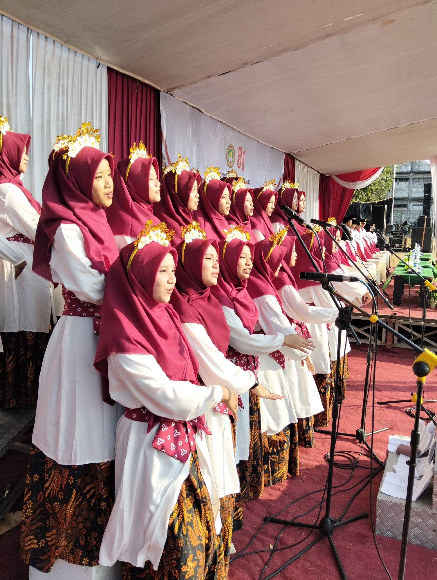 In Paciran, Indonesia, a group of young women, dressed in traditional attire, are performing on a stage. Each woman wears a deep red hijab and a white tunic with a patterned sash. Their outfits are adorned with golden headpieces featuring white flowers. They are standing in a line, facing forward, and appear to be singing, as indicated by their open mouths. Microphones are positioned in front of them, suggesting a musical performance or speech. The stage is covered in a red carpet, and a white banner with red accents hangs in the background, displaying the number "81" prominently. To the right of the stage, speakers and audio equipment are visible, along with a table holding water bottles and papers. The scene is illuminated by natural sunlight, casting shadows on the stage. The overall impression is of an outdoor cultural event or ceremony.