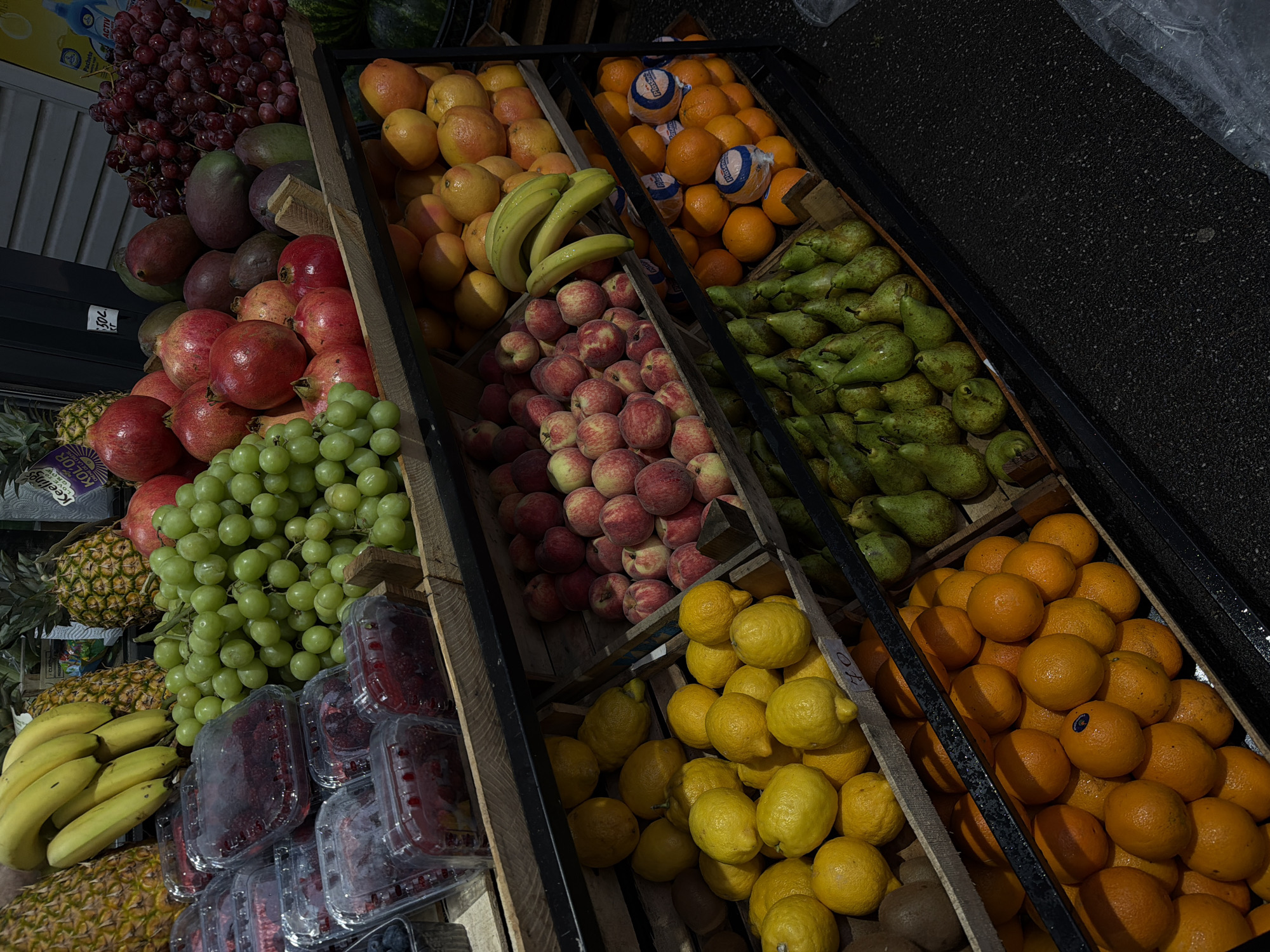 The image shows a fruit stand in an unspecified urban location. Wooden crates are filled with a variety of fruits, including red and green grapes, mangoes, pomegranates, pineapples, bananas, raspberries, blueberries, oranges, peaches, pears, and lemons. The scene is captured in natural lighting.