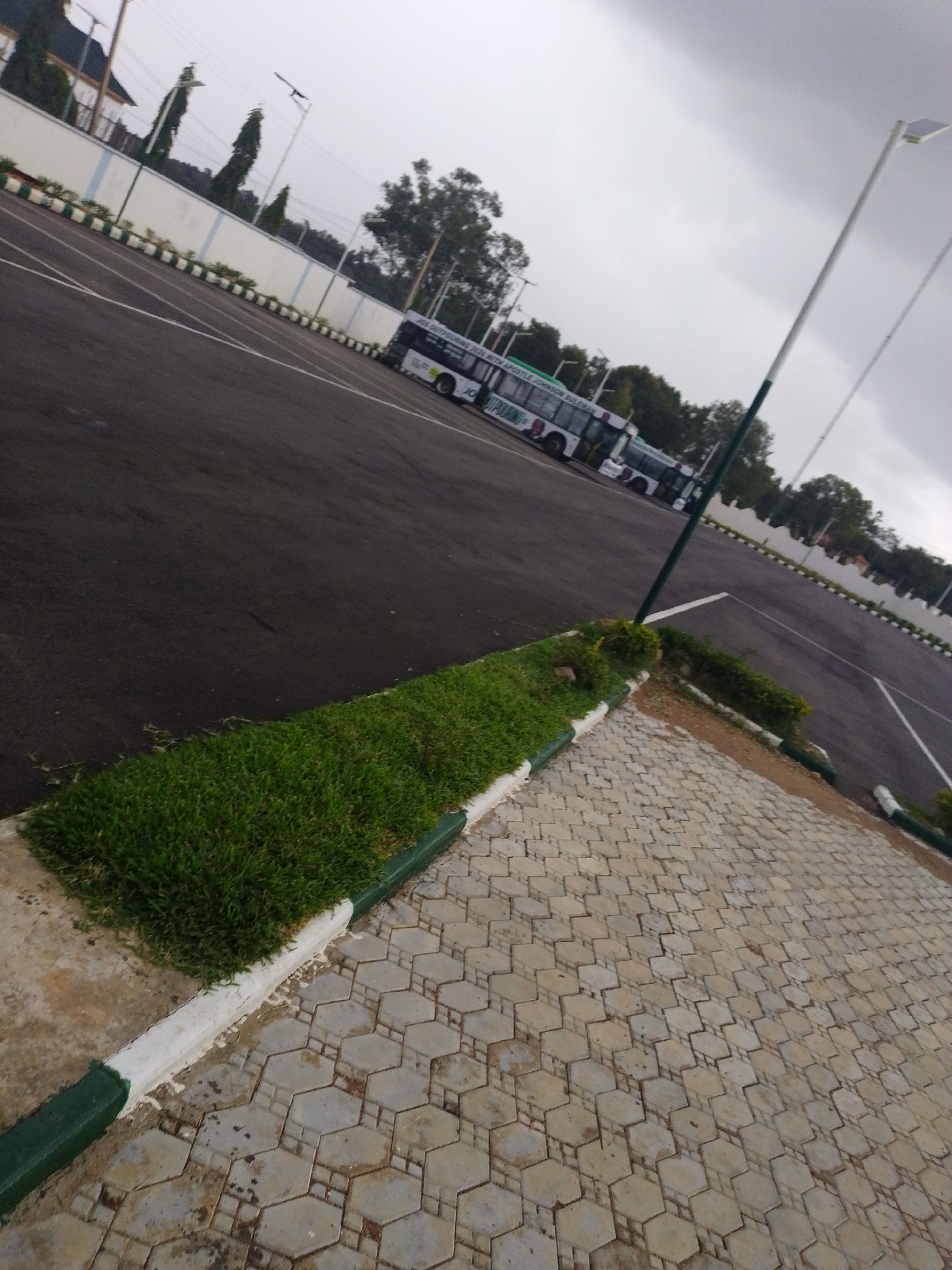 The image shows a large, paved parking lot with several white buses parked in the background. A strip of green grass and decorative border separates the parking lot from a hexagonal-tiled walkway in the foreground. White lines delineate parking spaces in the asphalt. Tall light poles are visible, and the sky is overcast. The setting appears to be an outdoor area, possibly a facility or event ground, with a wall and some trees in the distance. The image does not provide specific clues to definitively place it in Barikin Jirga Summa, Nigeria, but it depicts a typical urban or semi-urban parking and walkway environment.