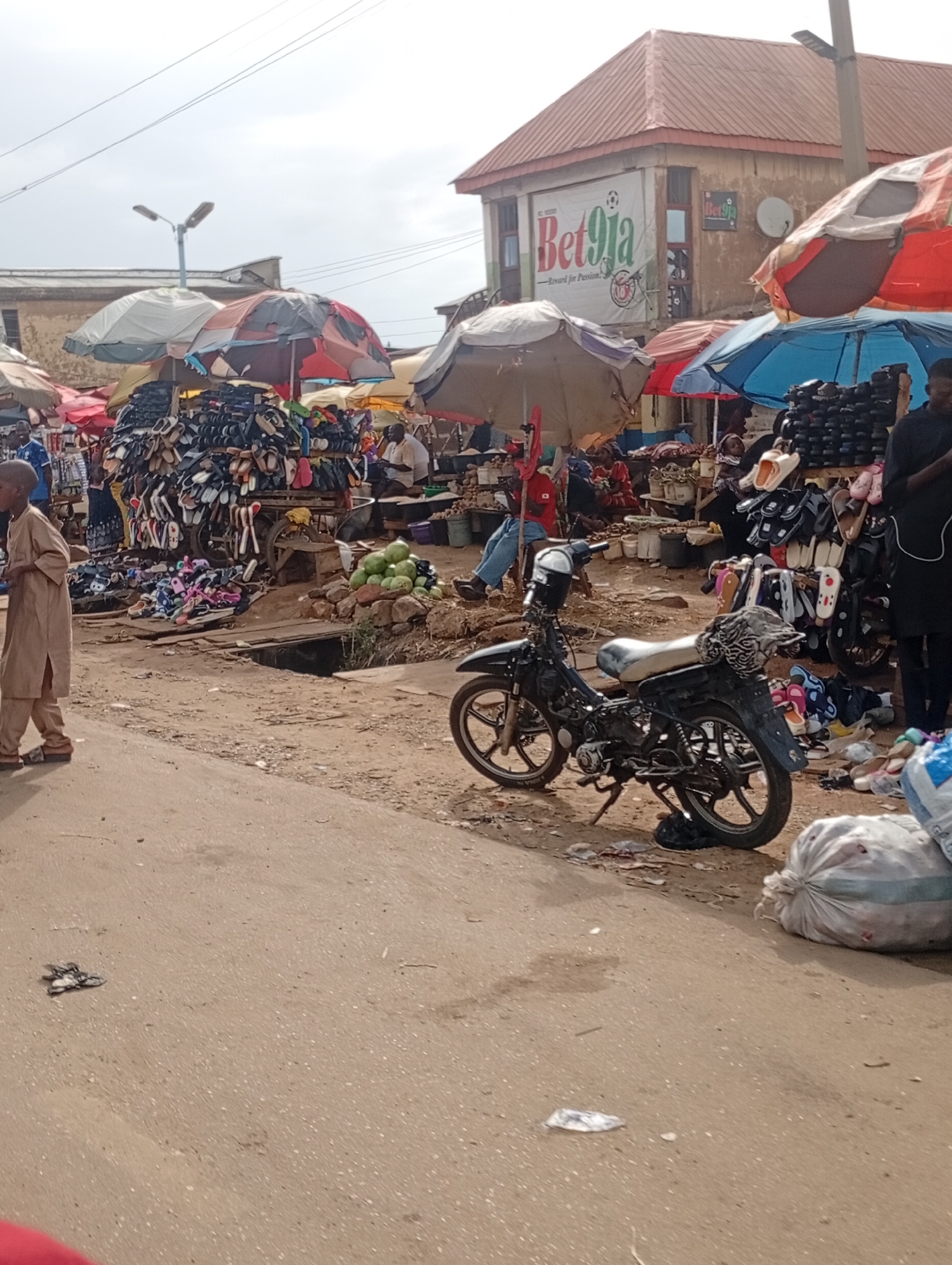 The image depicts a busy market scene in Rafin Par, Nigeria. In the foreground, a young boy dressed in a light brown traditional outfit walks from left to right. Beside him, a black motorcycle is parked, its kickstand down. In the background, various market stalls are set up under colorful umbrellas. One stall prominently displays a large quantity of footwear, including sandals and flip-flops, arranged on shelves and a cart. Another stall features a pile of green produce, possibly watermelons.
Several people are visible in the market. A man in a black outfit stands near the motorcycle, to the right. Other individuals are partially obscured by the stalls and umbrellas, engaged in shopping or selling. The overall atmosphere suggests an active marketplace, likely during daytime, given the bright lighting. The ground is dirt and pavement, indicative of an outdoor setting.
A notable detail is a sign on a building in the background that reads "Bet9ja" with accompanying graphics. This likely indicates a betting shop or advertising related to it. The weather appears to be overcast, with a light sky. The scene is typical of a bustling street market in Nigeria, with a mix of goods, people, and vehicles.