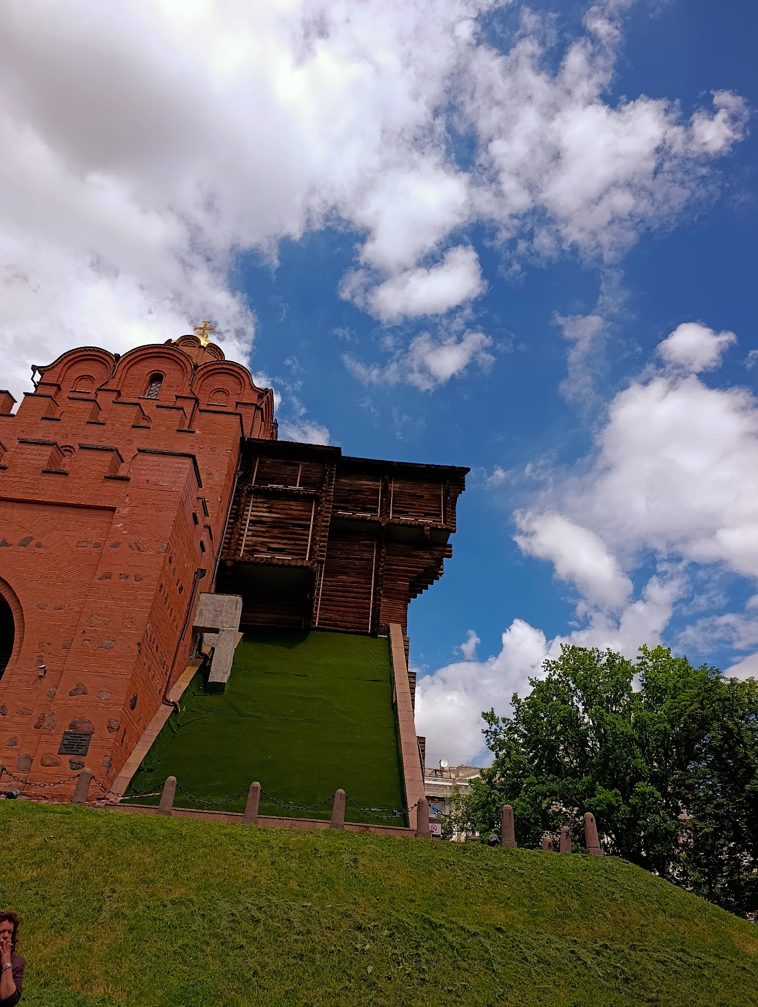 The image shows the Golden Gate of Kyiv, Ukraine. The gate is made of red brick and has a wooden structure attached to its side. A bright green lawn slopes up to the gate, and a chain fence with stone posts separates the lawn from the gate area. In the foreground, a woman with curly brown hair is visible on the far left, partially obscured and looking down. The sky is a vibrant blue with scattered white clouds. Green trees are visible to the right of the gate, and buildings can be seen in the distance. The perspective is from a low angle, looking up towards the gate and the sky.