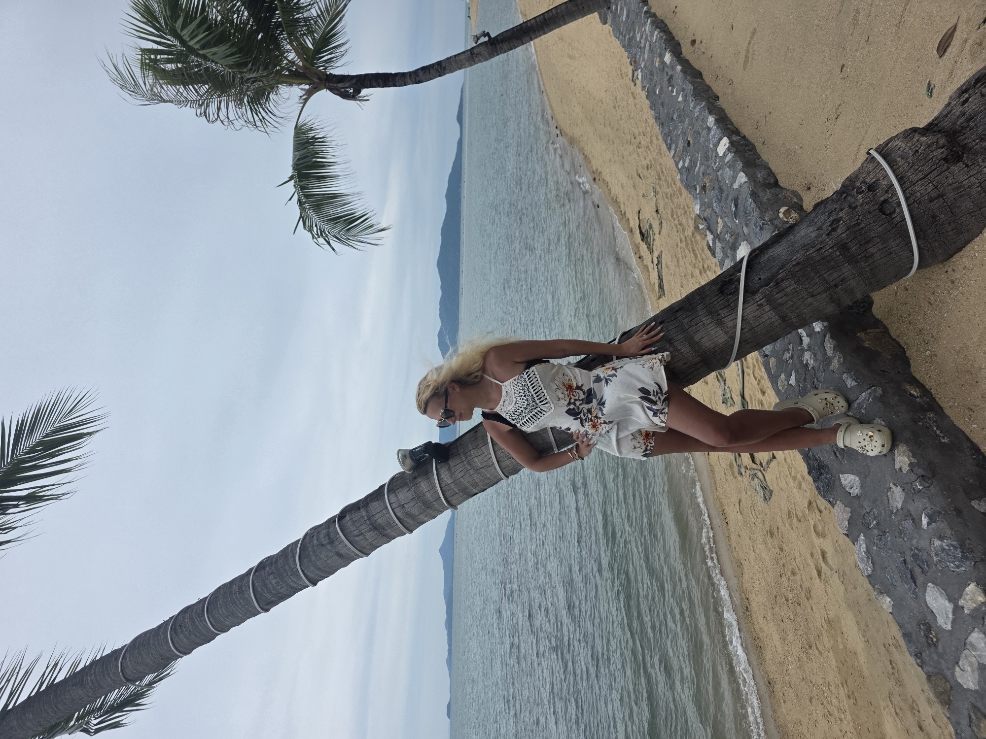A blonde woman stands on a fallen palm tree on a beach in Baan Bo Phut, Thailand. She is wearing a white floral dress and white clogs, with her hands on her hips and her head turned to the right. The sea is behind her, with distant islands and mountains visible on the horizon. The sky is overcast. A second palm tree, still upright, is to the right of the frame. A stone wall runs along the edge of the beach.