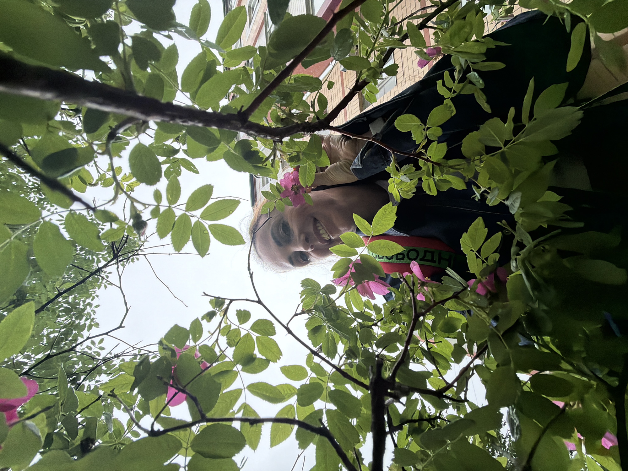 A woman smiles while looking up at the camera through green leaves and pink flowers. She is wearing a dark jacket over a lighter-colored shirt, and a red and green strap is visible across her chest. The background shows a building with beige siding and orange-brown brick accents. The shot is taken from a low angle, looking upwards through a plant.