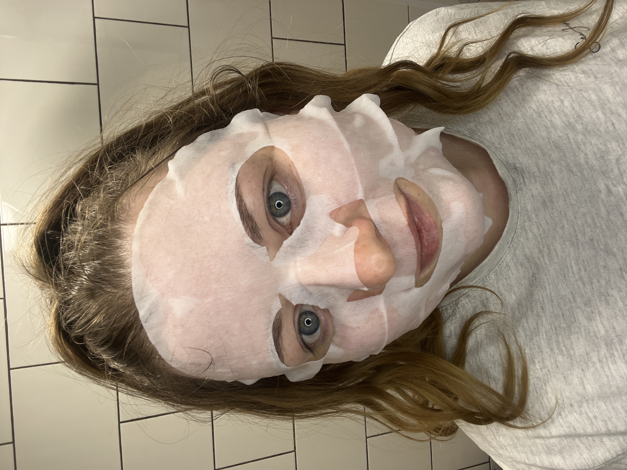 The image shows a person lying down with a facial sheet mask on. The person has light brown hair and blue eyes. They are wearing a grey t-shirt. The background appears to be a tiled wall. The location is not discernible from the image and cannot be identified as Раздолье, Russia.