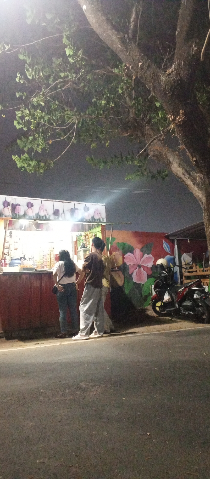 In the city of Madiun, Indonesia, at night, three individuals are standing by a food stall. The stall has a red base and is illuminated by overhead lights, displaying various items for sale. The backdrop of the stall is a mural of large pink flowers and green leaves. To the right of the stall, a red motorcycle is parked. The scene is captured from a low angle, with a large tree and its branches dominating the upper portion of the image against a dark, hazy sky. The ground in front of the stall is an asphalt road. The people are facing away from the camera, with two of them appearing to be customers and the third possibly an attendant.