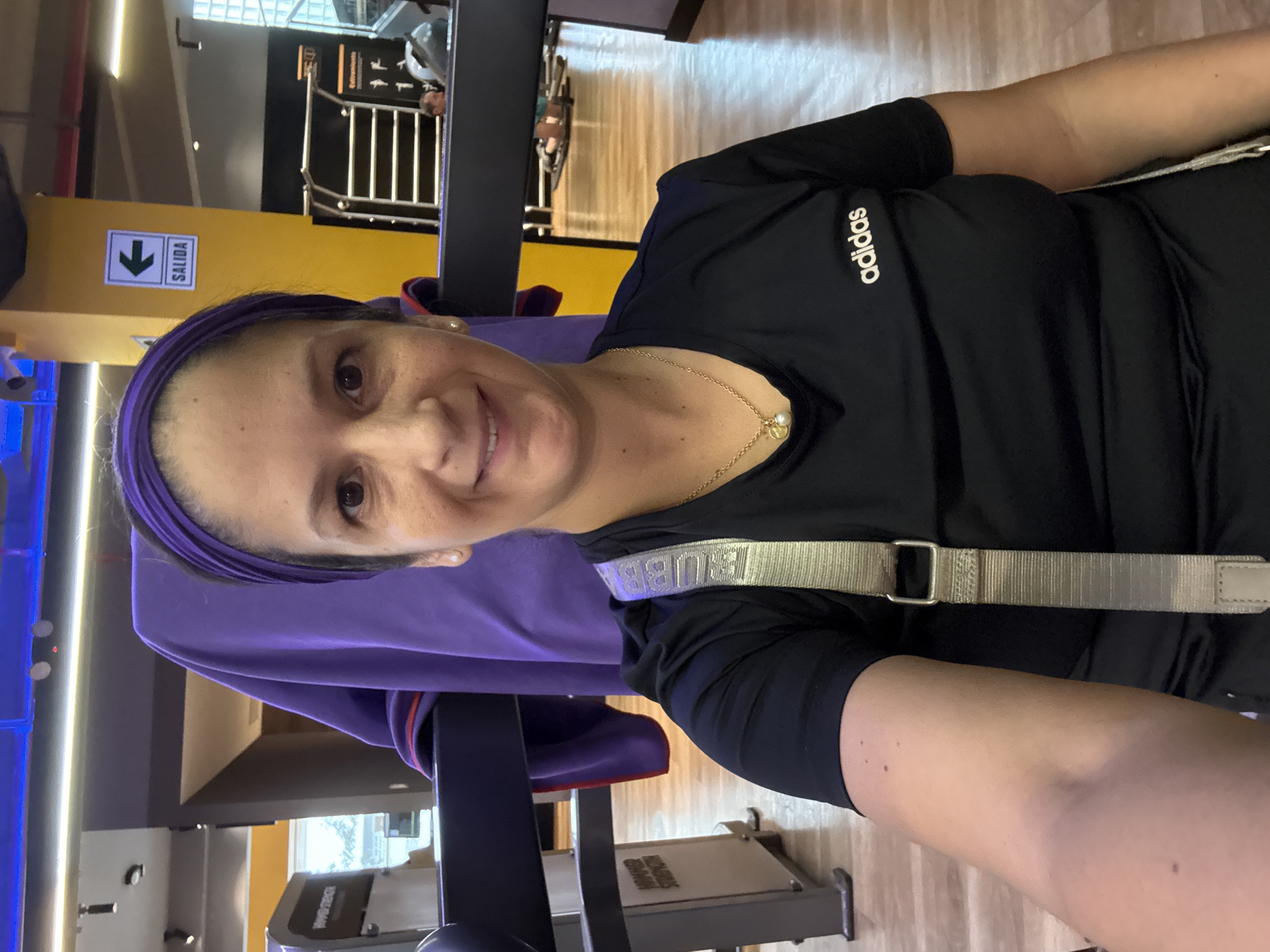 The image shows a woman in a gym, wearing a black Adidas t-shirt and a purple headband. She is smiling and looking at the camera. The setting appears to be a modern gym with workout equipment visible in the background. There's a sign with an arrow pointing down and the word "SALIDA" (exit in Spanish), suggesting this is a public space. The lighting suggests it's daytime. The woman's expression is happy and relaxed. She is also wearing a light-colored strap across her chest, possibly part of a bag or equipment.