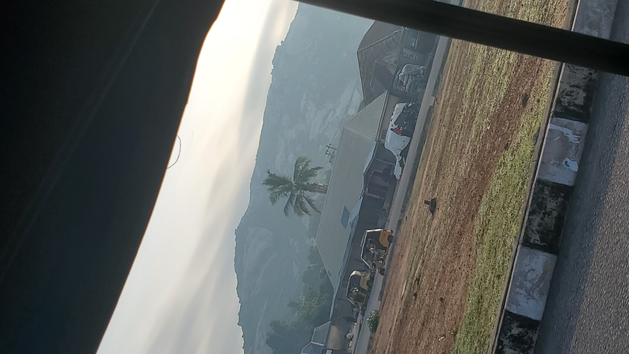 The image depicts a roadside scene in Jalingo, Nigeria, with a mountainous landscape in the background under a hazy sky. In the foreground, a paved road is visible, bordered by a low concrete barrier with alternating black and white painted sections, showing some wear and tear. Adjacent to the road is an area of dry, brown earth interspersed with patches of green grass. Several buildings with corrugated metal roofs are situated along the street. Parked or moving along the street are at least three yellow auto rickshaws, commonly known as 'Keke Napep'. One building appears to be a small shop or market stall. A single palm tree stands prominently in front of one of the buildings, with its fronds spread wide. The overall impression is of a typical urban or peri-urban environment in Nigeria, likely captured during daylight hours.