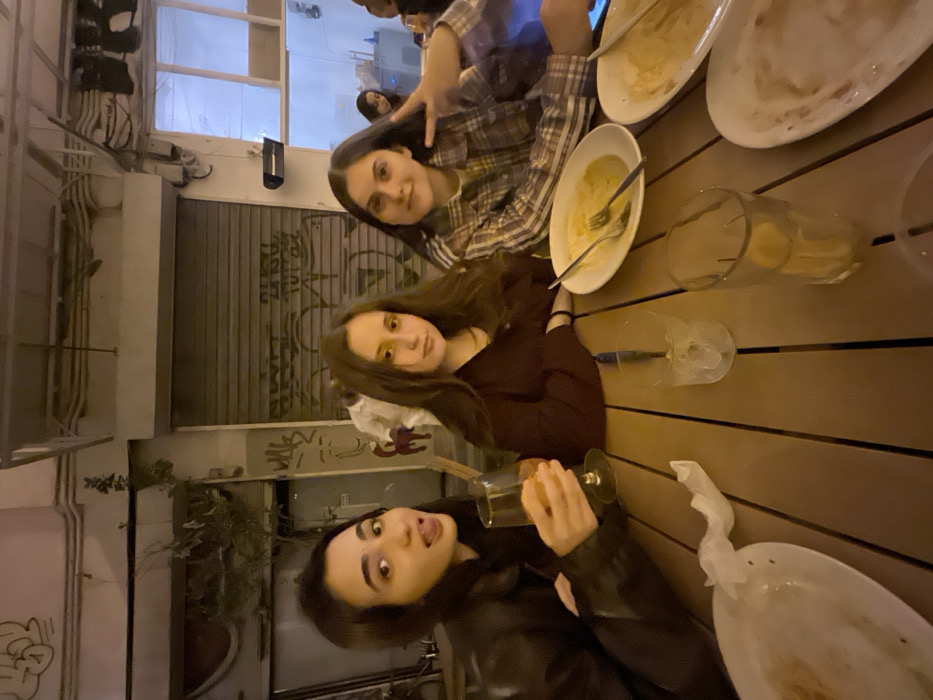 Three young women are at a table with empty plates and glasses. The woman at the top is making a peace sign and wearing a plaid shirt. The woman in the middle is wearing a dark red shirt. The woman at the bottom is wearing a black leather jacket and sticking out her tongue. The table is made of wood and there is a building in the background.