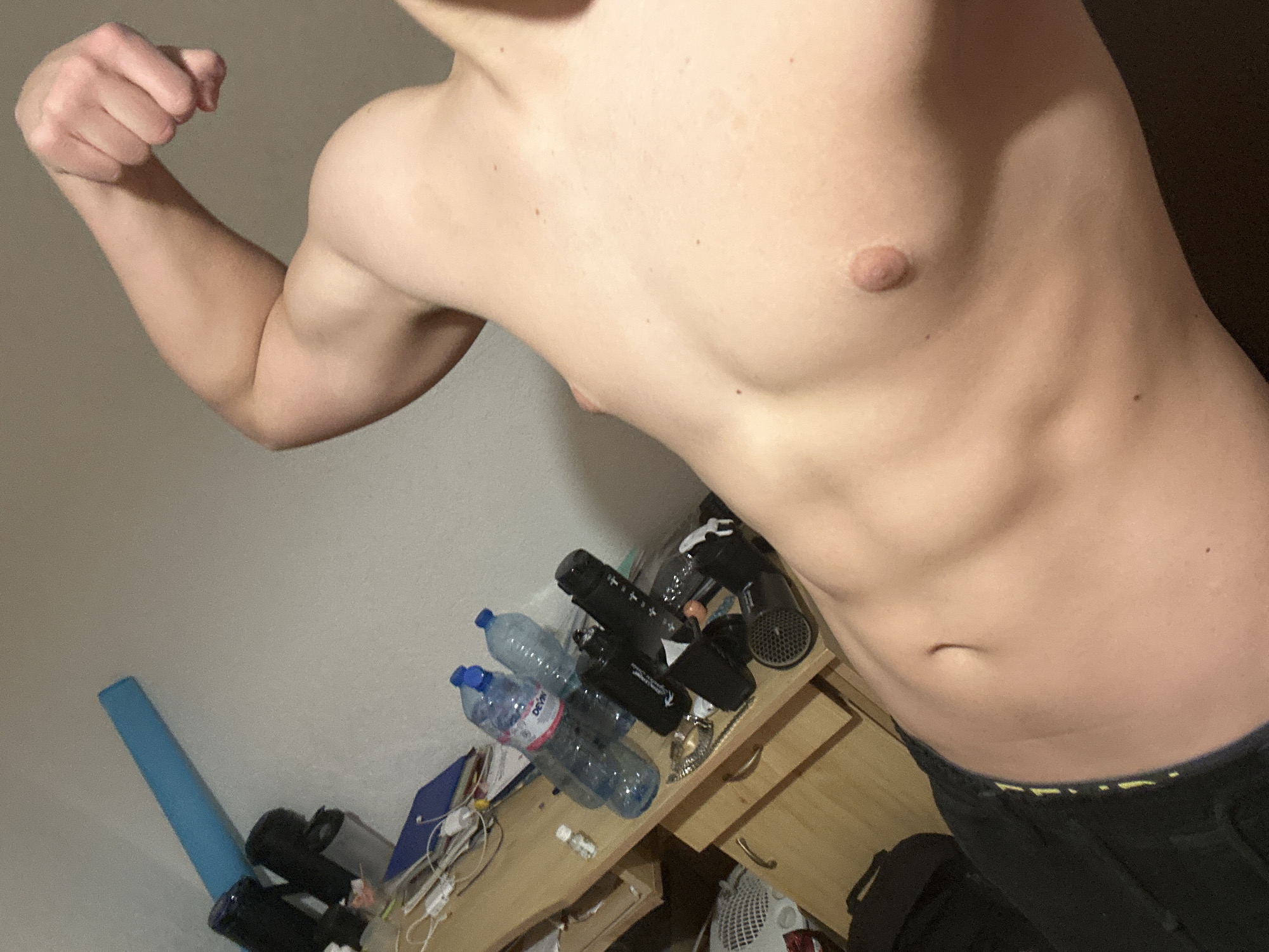 A shirtless person is flexing their right bicep. The person has a light complexion. The background includes a wall and a dresser with various objects, including water bottles, hair styling tools, and a blue foam roller. The location in the city of Zlatograd, Bulgaria, is unknown.