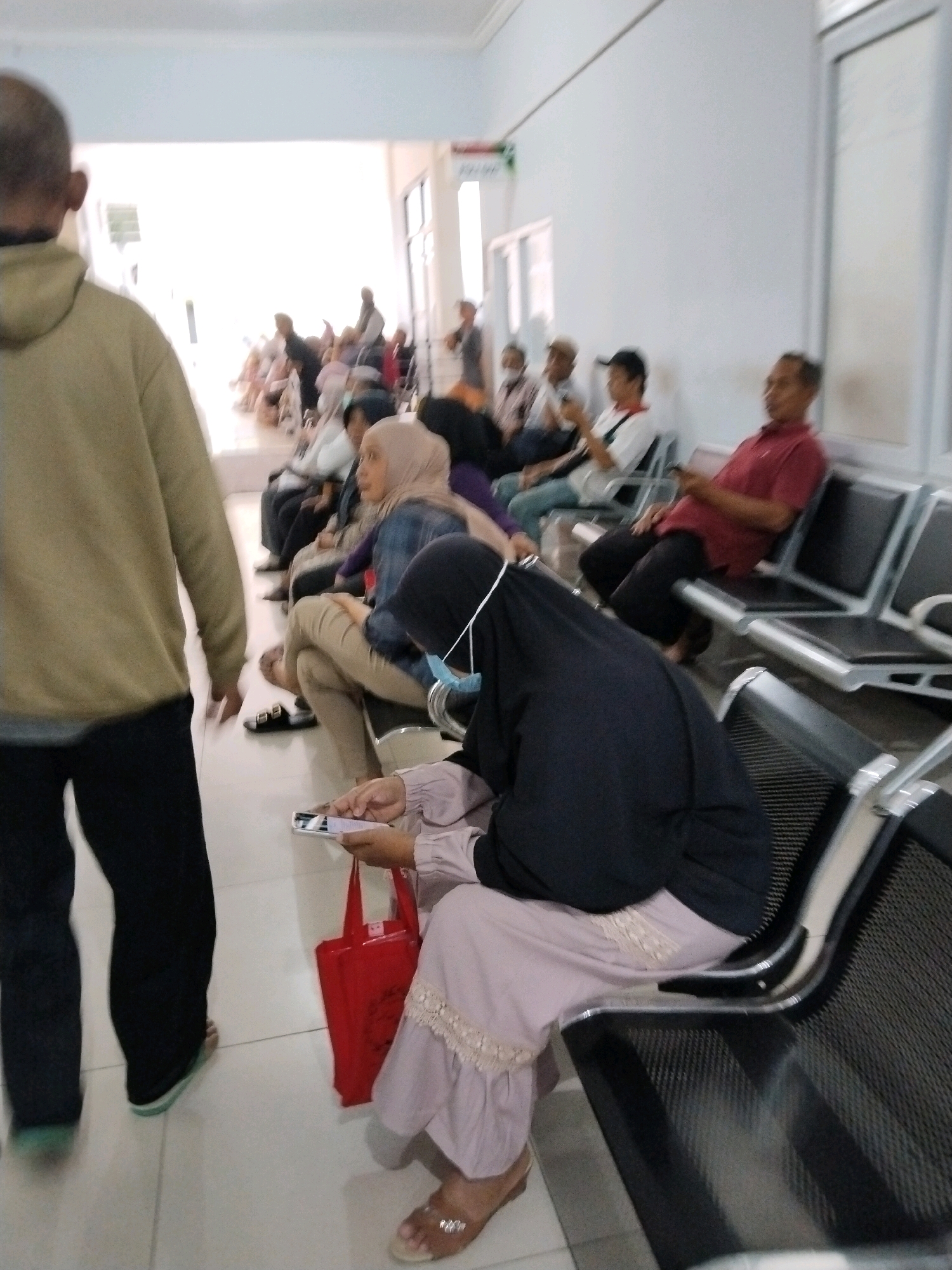 The image depicts a waiting area, possibly a clinic or government office, in the city of Trenggalek, Indonesia. Several people are seated on benches, facing forward or to the side, suggesting they are waiting for an appointment or service. In the foreground, a person wearing a black head covering and a light-colored dress is holding a smartphone and a red bag. Another person, partially visible, is standing in the left foreground, wearing a light brown hooded jacket and dark pants. The environment appears to be indoors, with white walls and a tiled floor. The lighting is bright, suggesting daylight. The overall atmosphere is one of quiet anticipation, with individuals engrossed in their own activities or observing their surroundings.