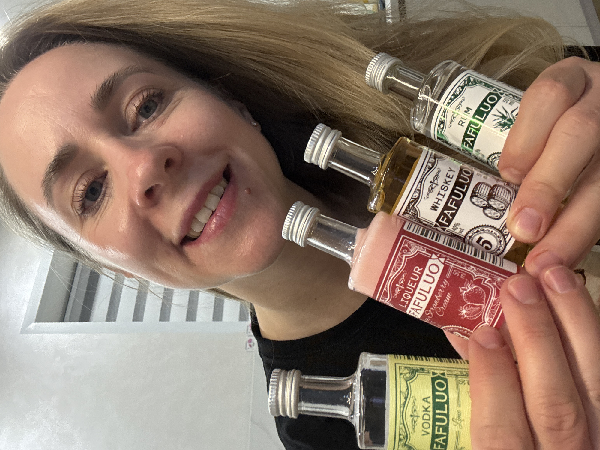 A Caucasian woman is holding four miniature bottles of liquor with labels that read "FAFULUO" in an indoor setting. The bottles are labeled as: Rum, Whiskey, Liqueur Strawberry Cream, and Vodka Lime. The woman is fair-skinned with blonde hair. She is smiling and looking at the camera. The background is slightly blurred and out of focus. The image does not provide enough information to determine the location as Chaikovsky, Russia.