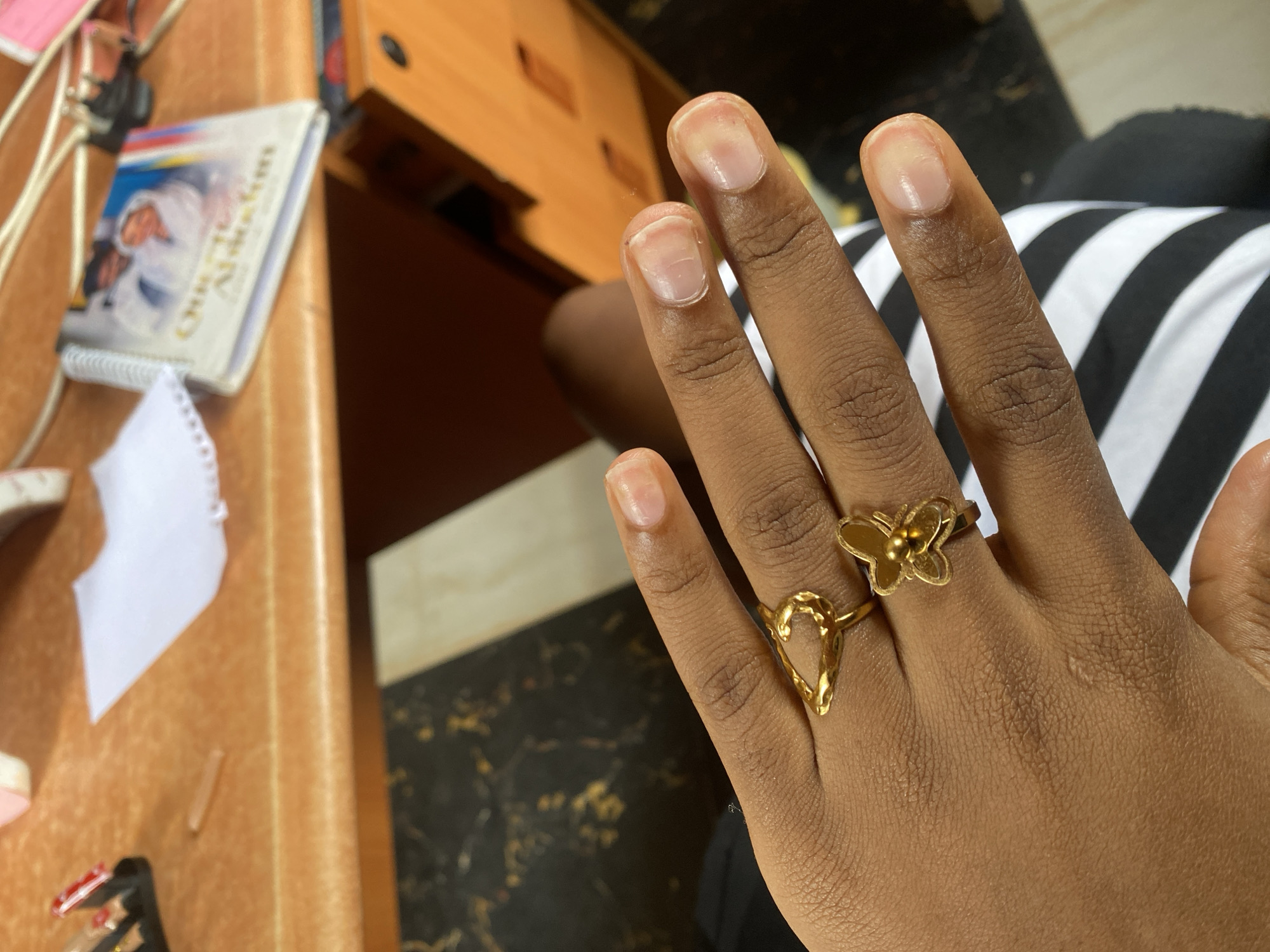 A close-up shot focuses on the left hand of an individual wearing two gold rings, one shaped like a heart outline and the other like a butterfly. The hand is resting on a black and white striped garment. In the background, on a light brown wooden surface, a small, white notepad is visible next to a spiral-bound booklet with an image of two people on its cover. The overall environment suggests an indoor setting, possibly a desk or table, with the surface exhibiting a subtle wood grain texture. The lighting is diffused, creating soft shadows. There are no identifiable landmarks or contextual clues within the image to pinpoint its location in Jos, Nigeria.