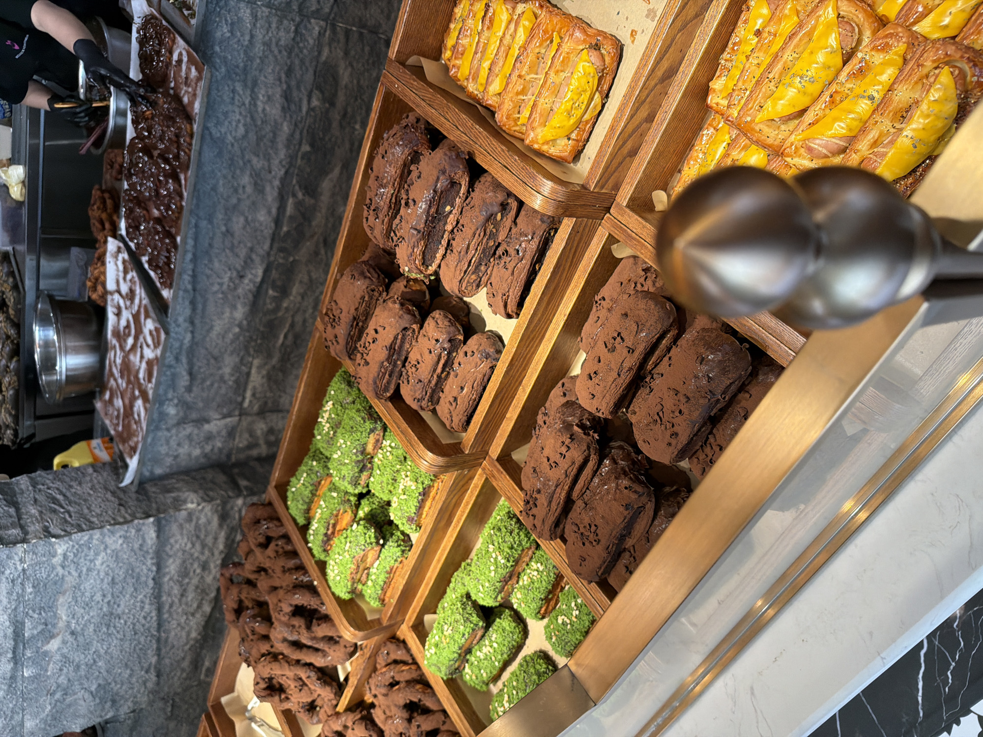 The image shows a bakery display case filled with various pastries. The case is made of wood and is divided into several sections. In the top section are pastries with yellow toppings. Below these are pastries covered in chocolate powder. Further down are pastries topped with green sprinkles, with chocolate pastries and fried dough twists below. A person wearing black gloves and a black shirt is visible in the upper left corner, appearing to be handling the pastries. The photograph appears to have been taken from an elevated angle. The scene is in a high-end bakery, potentially located in the Chongqing High-tech Zone, China.