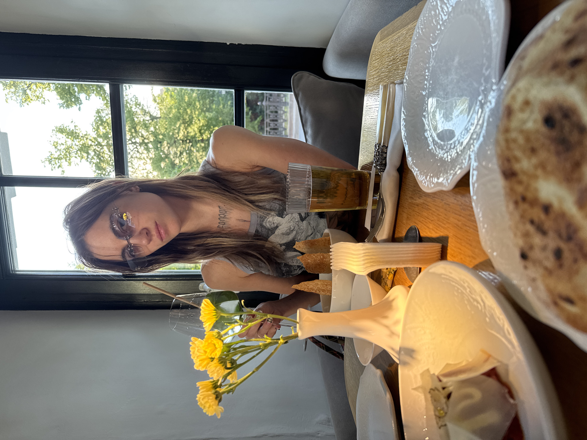 A young woman with long, light brown hair and sunglasses is seated at a wooden table in what appears to be a restaurant or cafe. She wears a casual grey top with a pattern and has a tattoo with partially obscured text on her left arm. She looks directly at the camera with a neutral expression.
Behind her, a large window with a black frame reveals a bright outdoor scene with green trees and foliage, indicating daytime. The table is set with white plates, decorative cutlery, and a white napkin. A glass containing an amber-colored drink with a straw is on the table, along with a white vase holding vibrant yellow flowers, possibly chrysanthemums. Other dining elements include a bowl of what looks like toasted flatbread or chips, and additional food items on a plate, all suggesting a meal is in progress or about to be served. A grey upholstered chair or booth is visible behind the woman. The setting is well-lit by natural light from the window.