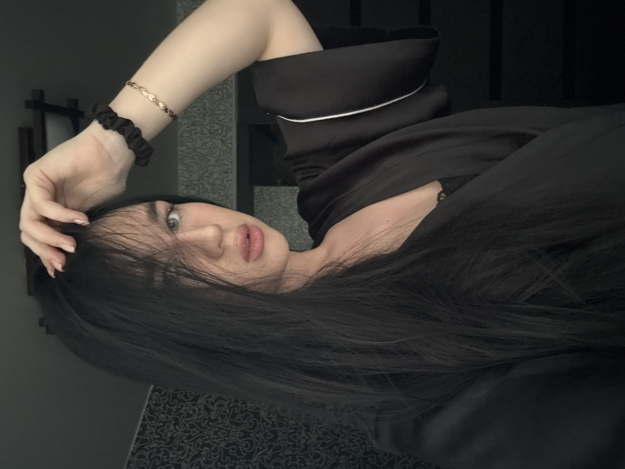 The image is a close-up selfie of a young woman with long, dark hair, fair skin, and light-colored eyes. She is wearing a black robe with white trim and a black scrunchie on her right wrist, along with a gold bracelet. Her right hand is raised to her head, and her hair is partially covering her face. Her lips are slightly parted. The background appears to be an indoor setting with textured wallpaper and a dark, minimalist decor. There are no discernible landmarks or elements that would indicate a specific location within the city of Novosibirsk, Russia.