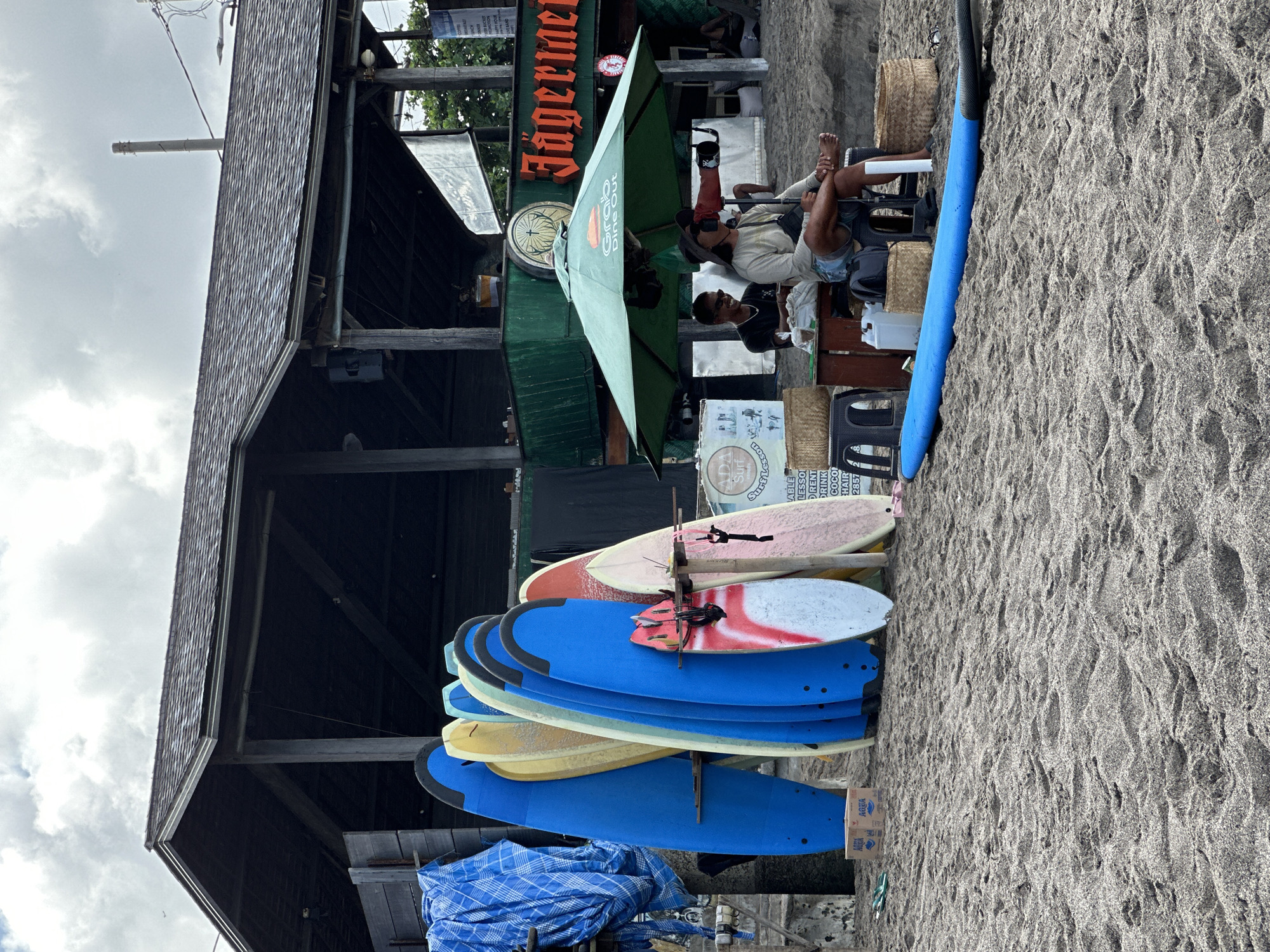 The image shows a beachfront scene in North Kuta, Indonesia. A rack of surfboards of various colors, mostly blue, leans against a wooden structure with a shingled roof. Behind the surfboards, a Jägermeister-branded outdoor bar area is visible, with a green umbrella shading patrons. People are seated at tables, one with their feet up, seemingly relaxing. The ground is composed of dark, sandy soil.
