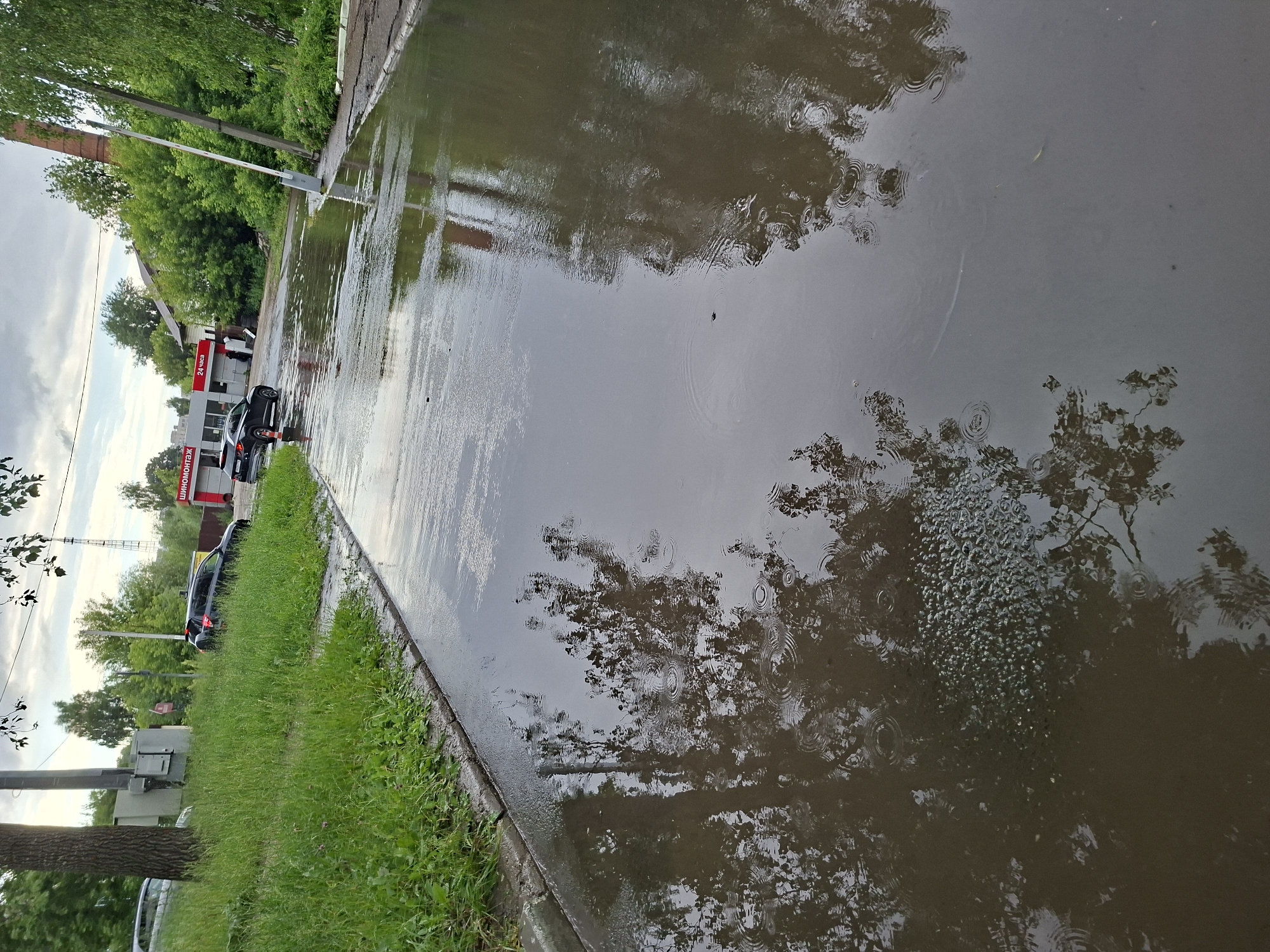 The image depicts a flooded street in Kazan, Russia, likely after heavy rainfall. A large puddle occupies the majority of the foreground, reflecting the surrounding trees and buildings. The water level is high enough to cover a significant portion of the asphalt road, with ripples visible on its surface, suggesting continued or recent precipitation.
On the left side of the image, a grassy embankment runs alongside the road, with a tall tree trunk and a utility box. A dark-colored SUV is parked on the side of the road, near a building with a red and white sign that reads "ШИНОМОНТАЖ 24 ЧАСА", indicating a tire service operating 24 hours. Another dark-colored car is further down the road, partially submerged in the floodwater.
Further in the background, more trees and buildings are visible, suggesting a residential or commercial area. A white boom barrier, commonly used to control vehicle access, is also present on the right side of the road. The sky is overcast, with a dull, cloudy appearance. The overall scene conveys a sense of disruption caused by the flooding, impacting traffic and daily activities.