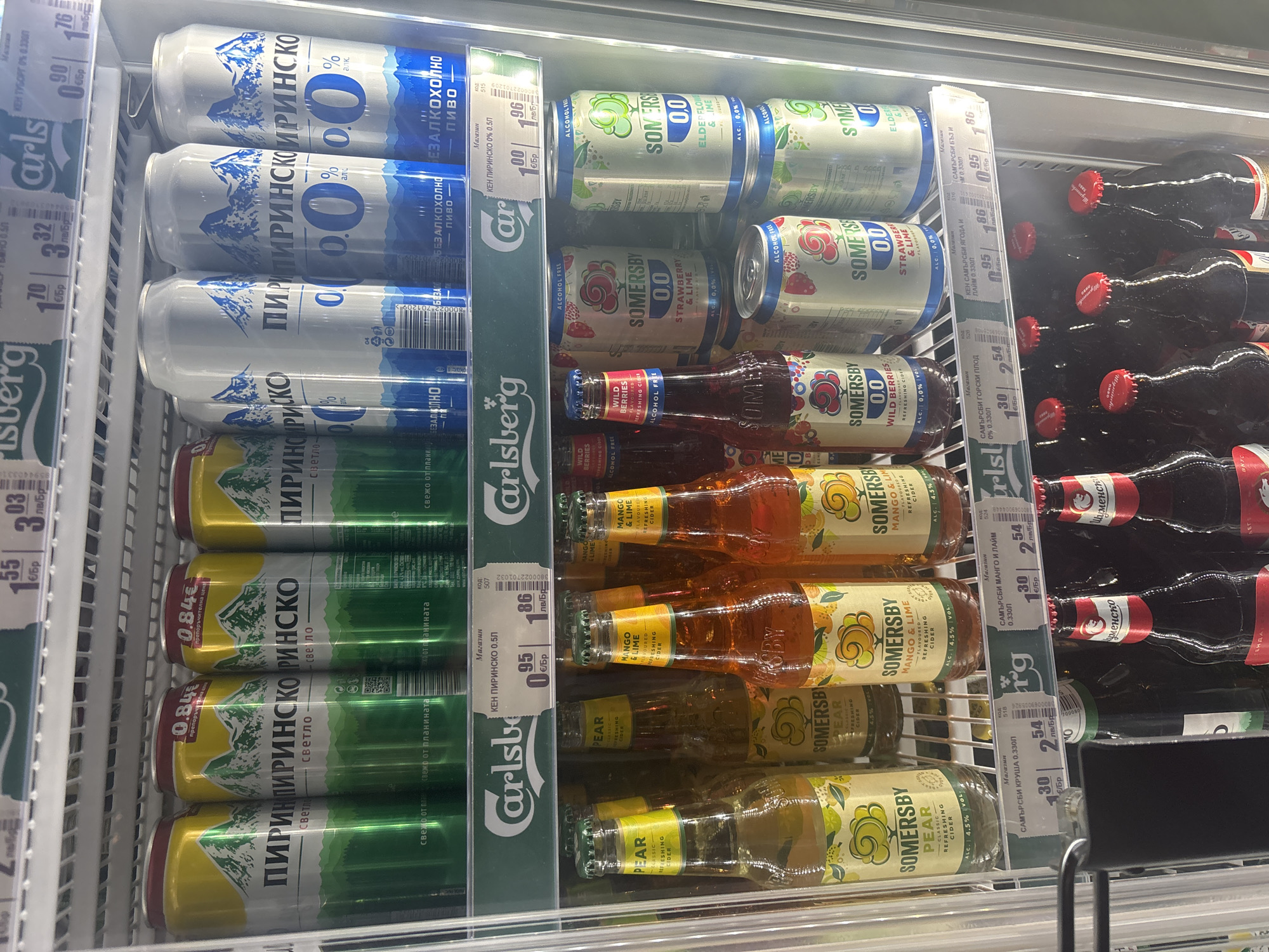 The image shows a refrigerated display case, likely in a store in Plovdiv, Bulgaria, featuring various alcoholic and non-alcoholic beverages. Cans of "Пиринско" beer (both regular and 0% alcohol) are stacked on the left side. Next to them are cans and bottles of "Somersby" cider in different flavors such as elderflower & lime, strawberry & lime, wild berries, mango & lime, and pear. Price tags indicate prices in both Bulgarian lev (лв) and euro (€). The case appears to be well-stocked and organized.