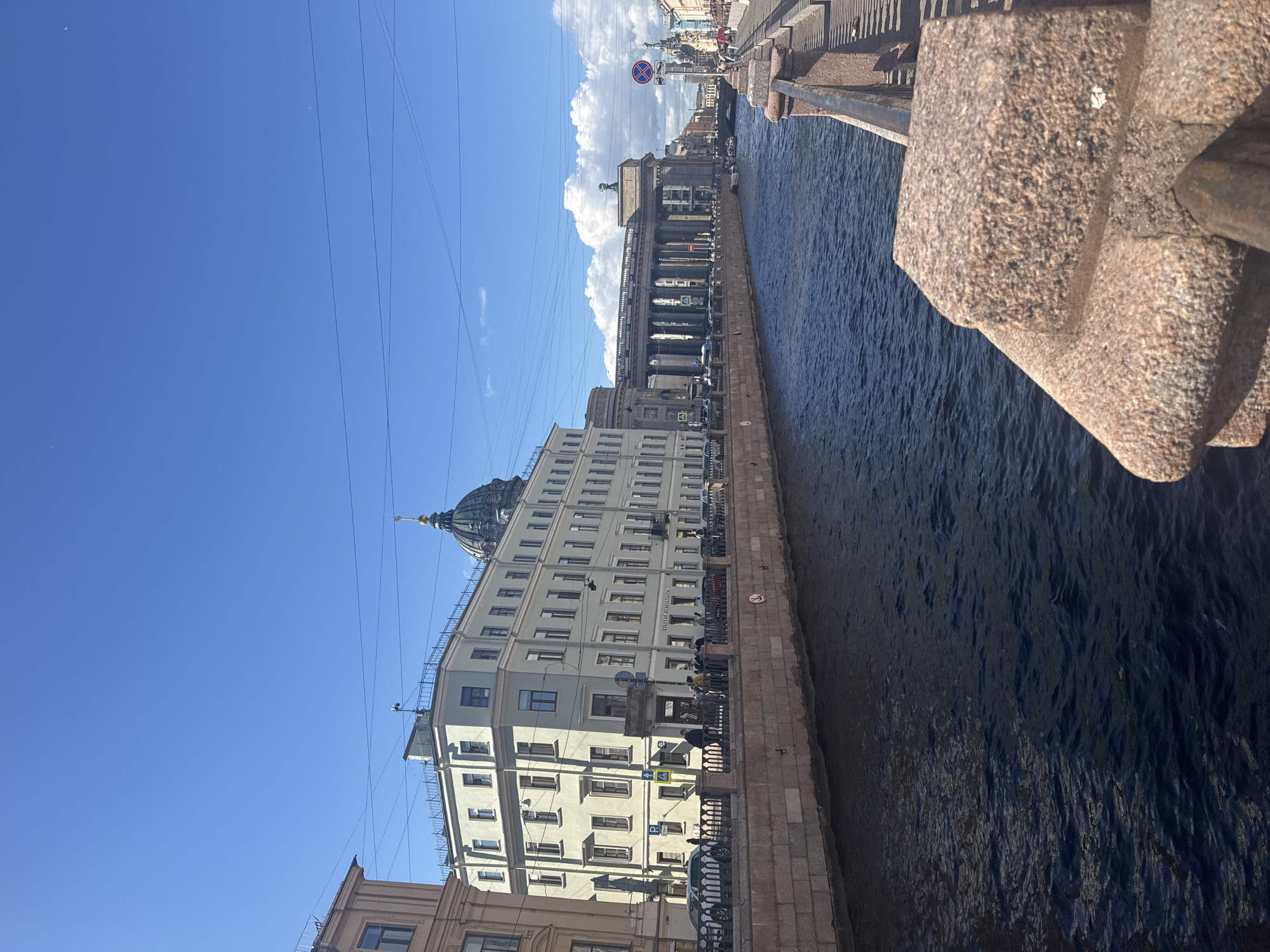 The image shows a wide canal in Saint Petersburg, Russia, with buildings lining its banks. On the left, a light-colored building with many windows and a prominent dome is visible. Across the canal, a grand neoclassical building with a long colonnade and sculptures is the dominant feature. The canal itself is dark blue with ripples, and stone embankments line both sides. A walkway with a railing is visible on the right bank, where a few people are walking. The sky is clear blue with scattered white clouds, and power lines are strung across it. The overall impression is a sunny day in a historic city center.