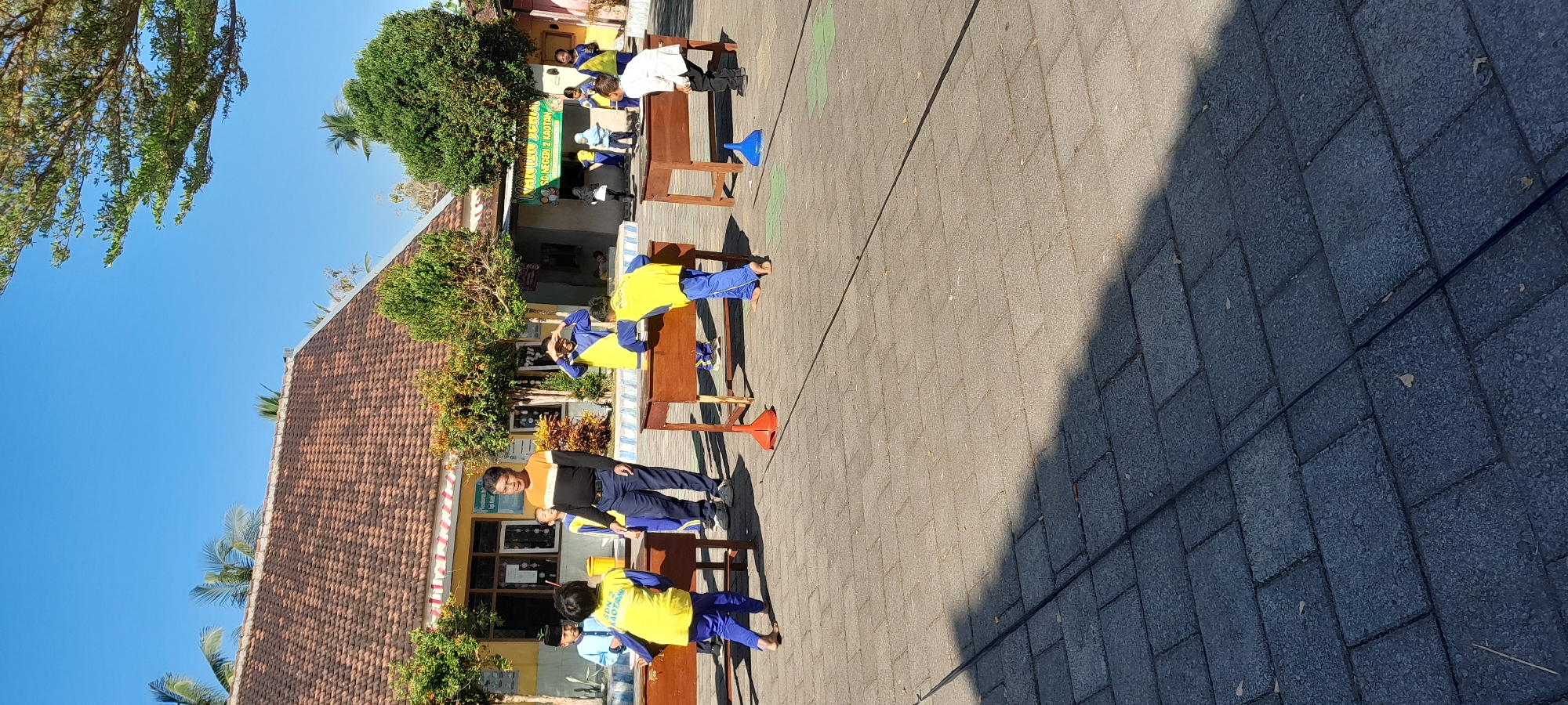 The image depicts a sunny outdoor scene, likely at a school in Rogojampi, Indonesia, given the presence of children in school uniforms and the building's architecture. Several young children, mostly wearing blue pants and yellow shirts with the inscription "SDN 2 KAOTAN," are engaged in an activity around wooden desks. Some children are bent over the desks, while others are standing or looking on. An adult male, wearing a black and orange shirt and dark pants, stands in the center of the frame, observing the children. The ground is paved with grey rectangular tiles. There are trees and buildings in the background, consistent with a school environment. The overall atmosphere appears to be one of a supervised outdoor educational or recreational event.