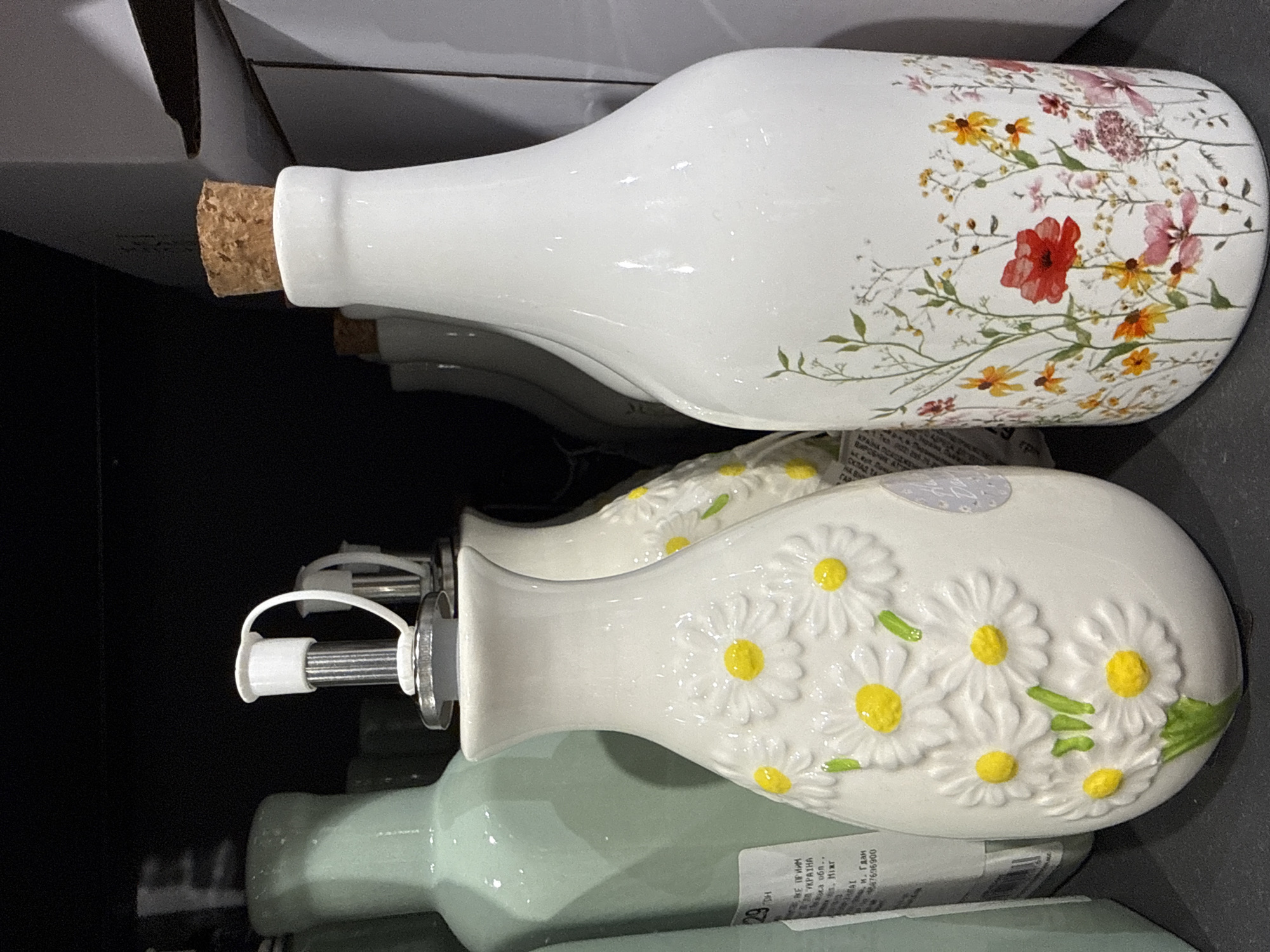 This media file features a close-up, top-down view of several decorative ceramic bottles, likely for oil or vinegar, arranged on a shelf. The primary subjects are the bottles themselves, two of which are in focus.
The topmost bottle is white with a delicate, colorful floral design featuring red, pink, yellow, and orange wildflowers and green stems. It has a cork stopper. Below this, a second bottle, also white, is adorned with raised white daisy-like flowers with yellow centers and green leaves. This bottle has a metal pour spout with a white plastic cap. The spout and cap assembly is clearly visible. Partially visible beneath the daisy bottle is a third bottle, a pale green color, which appears to be of a similar design but is out of focus.
The background is dark, suggesting an indoor setting, possibly a store display or a home shelf. The lighting is bright, highlighting the glossy finish of the ceramic bottles.
Visible text on a label attached to the green bottle reads: "КРАЇНА ВИРОБНИК УКРАЇНА" (Country of Origin: Ukraine) and other details in Ukrainian, including what appears to be product information and possibly a price or barcode. Another label with Cyrillic text is also partially visible on the green bottle's packaging.
The scene is static, with no discernible activity taking place beyond the display of the items. The overall impression is one of a retail or decorative setting showcasing kitchenware or home decor items. The location is identified as Odesa, Ukraine.