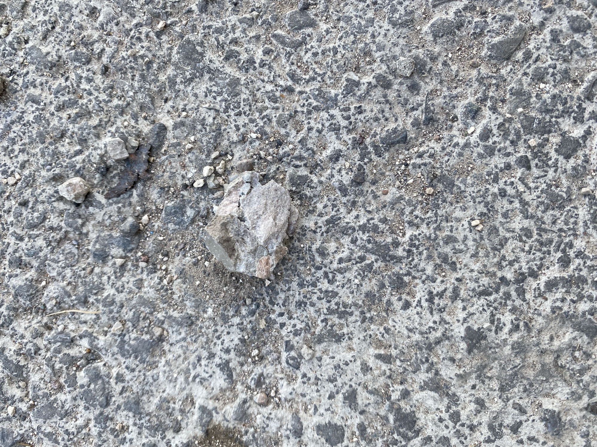 The image displays a close-up, overhead view of cracked and weathered asphalt or concrete, likely a section of a road or pavement. The surface is a mottled mix of dark gray and lighter gray tones, interspersed with small pebbles and gritty debris. A single, larger piece of light-colored rock or concrete debris is prominently featured slightly left of center, appearing rough and fractured. The texture of the ground is rough and uneven, with visible indentations and protrusions. The lighting suggests it is daytime, with no strong shadows indicating a specific time. The overall impression is of a mundane, urban or semi-urban outdoor surface. No people, animals, or any other objects besides the debris are visible. No text is present in the image. The location context of Yerevan, Armenia, does not provide any immediate visual cues within the image itself, as the scene is generic.