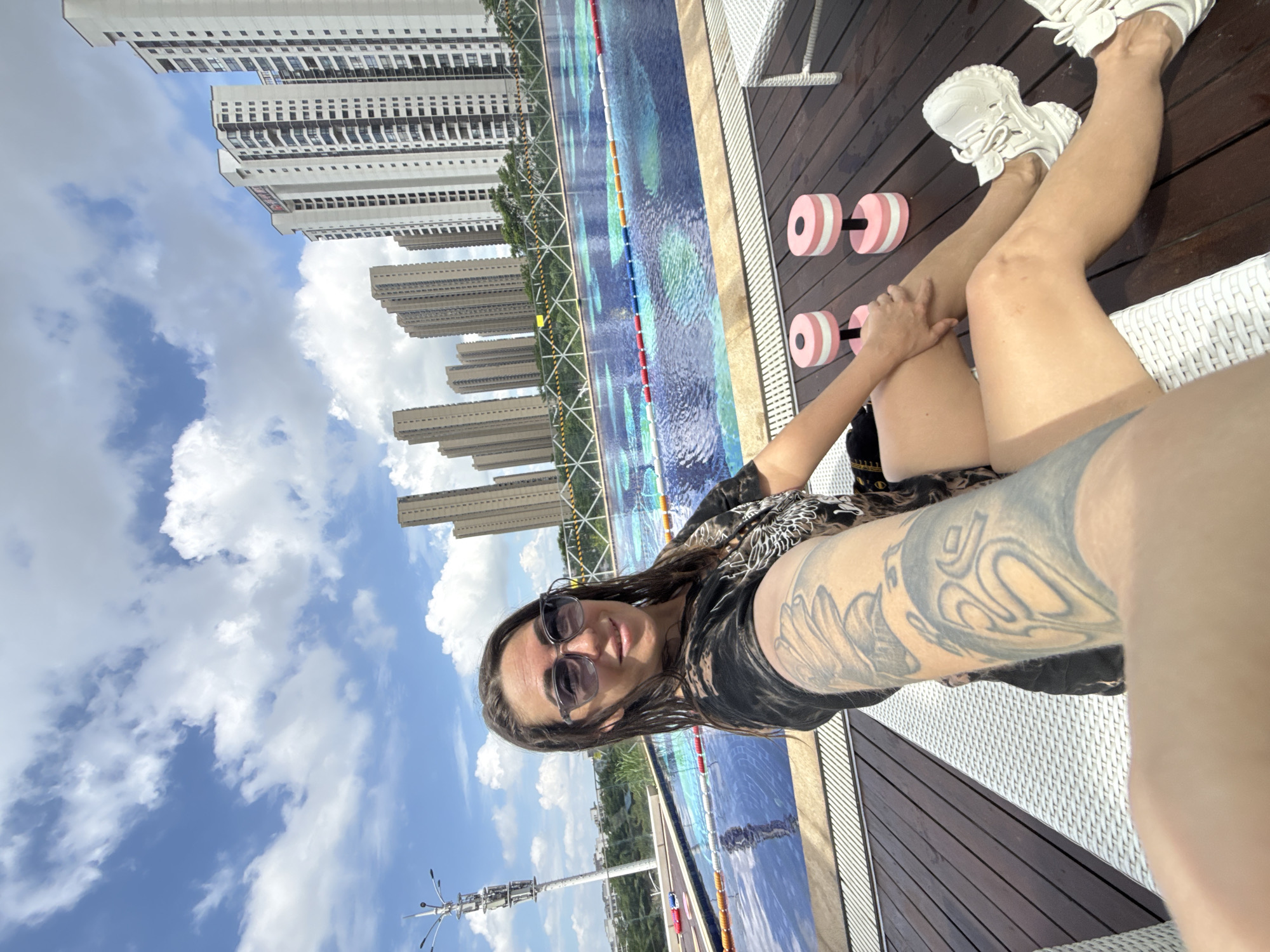 A woman with wet hair and sunglasses is taking a selfie on a wooden deck next to a swimming pool. She is wearing a black t-shirt with a tiger graphic and white sneakers. Two pink and white dumbbells are on the deck next to her. In the background, there is a large swimming pool with lane markers and several tall apartment buildings. The sky is blue with white clouds. The scene appears to be in a residential complex in Huizhou, China, likely a rooftop pool area based on the height of the buildings and the view.