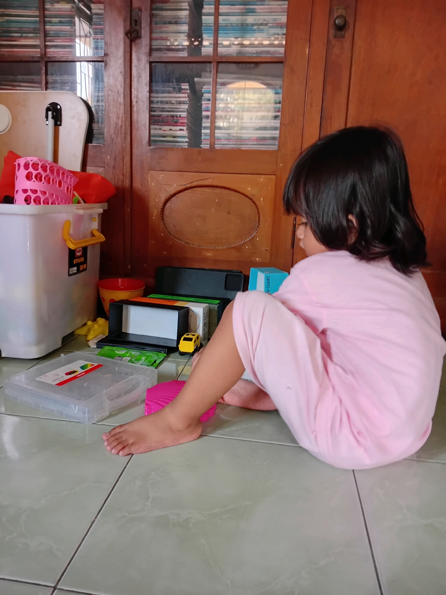 A young child, appearing to be of Southeast Asian descent with dark hair, is seated on a tiled floor in a room in Gebang, Indonesia. The child is wearing a light pink outfit and is facing away from the camera, looking towards a wooden cabinet with glass doors. Various objects are scattered around the child on the floor, including a white plastic storage bin, a pink basket, a red bucket, a yellow toy car, and several boxes and containers. The cabinet in the background is filled with what appear to be CDs or DVDs. The overall environment suggests a domestic setting, possibly a play area.