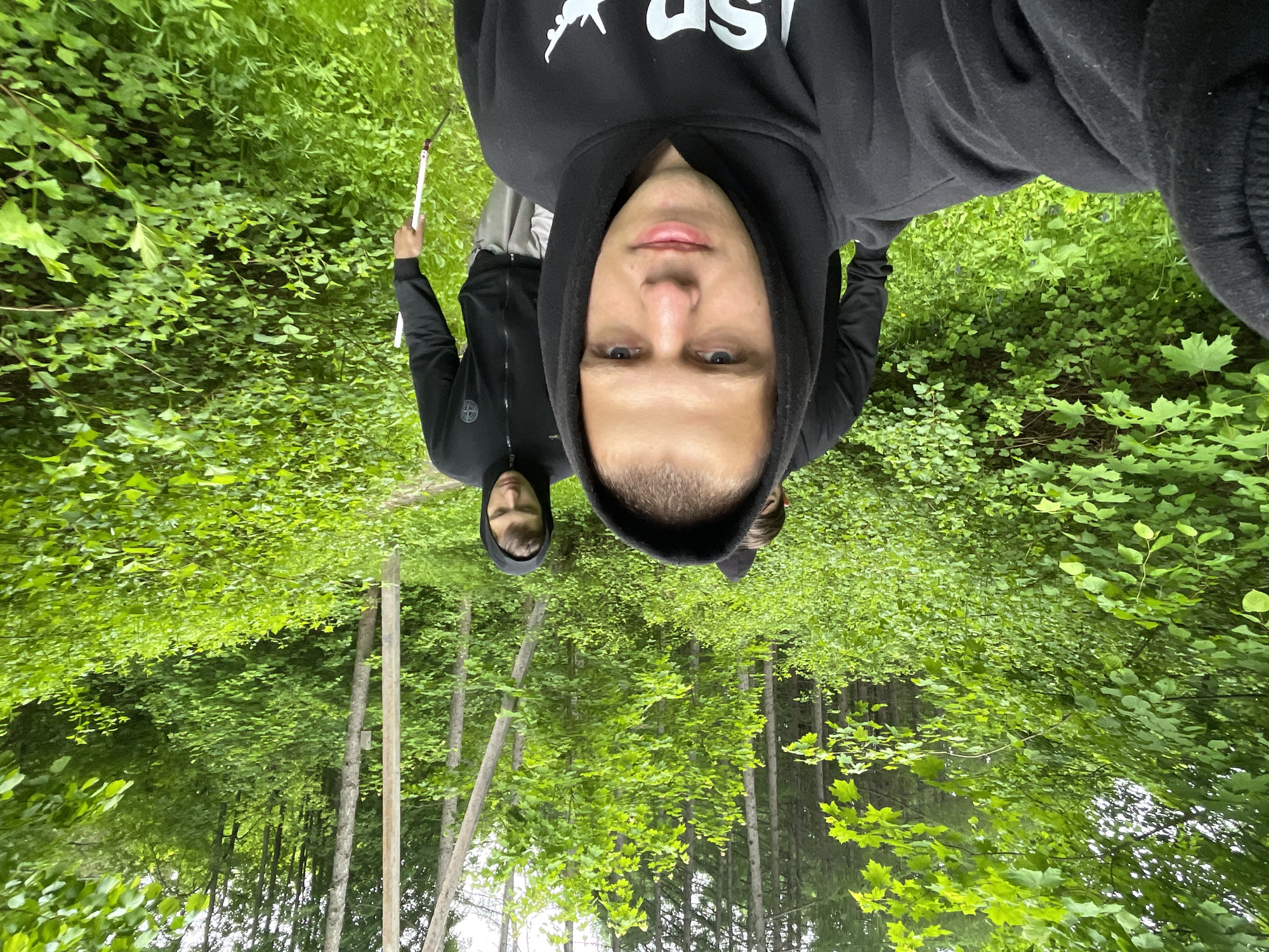 The image, correctly oriented, depicts two young men taking a selfie in a vibrant green forest or dense woodland, likely in Красные Баки, Russia. The time of day appears to be bright daytime, possibly under an overcast sky, as suggested by the even natural lighting.
The subject in the foreground is a young man, centrally positioned, wearing a black hooded sweatshirt with the hood up. His face is clearly visible, with light eyes and a neutral expression, looking directly into the camera, which he seems to be holding. On the chest of his hoodie, partially visible white text, likely "SP", and a white graphic resembling a stylized animal are present.
Behind him and slightly to the left, a second young man is visible, also wearing a black hooded top with the hood up. Only part of his face is seen, looking forward. He holds a long, slender, light-colored stick or pole with a dark tip in his right hand. A circular logo, consistent with the Stone Island compass badge, is visible on the left sleeve of his jacket.
The setting is characterized by an abundance of lush green foliage. The background features numerous tall, thin tree trunks reaching upwards, topped by a dense canopy of bright green leaves. The ground and mid-ground are covered in various green plants and undergrowth, including some leaves that resemble maple. The scene suggests a casual outing or exploration in a natural environment.