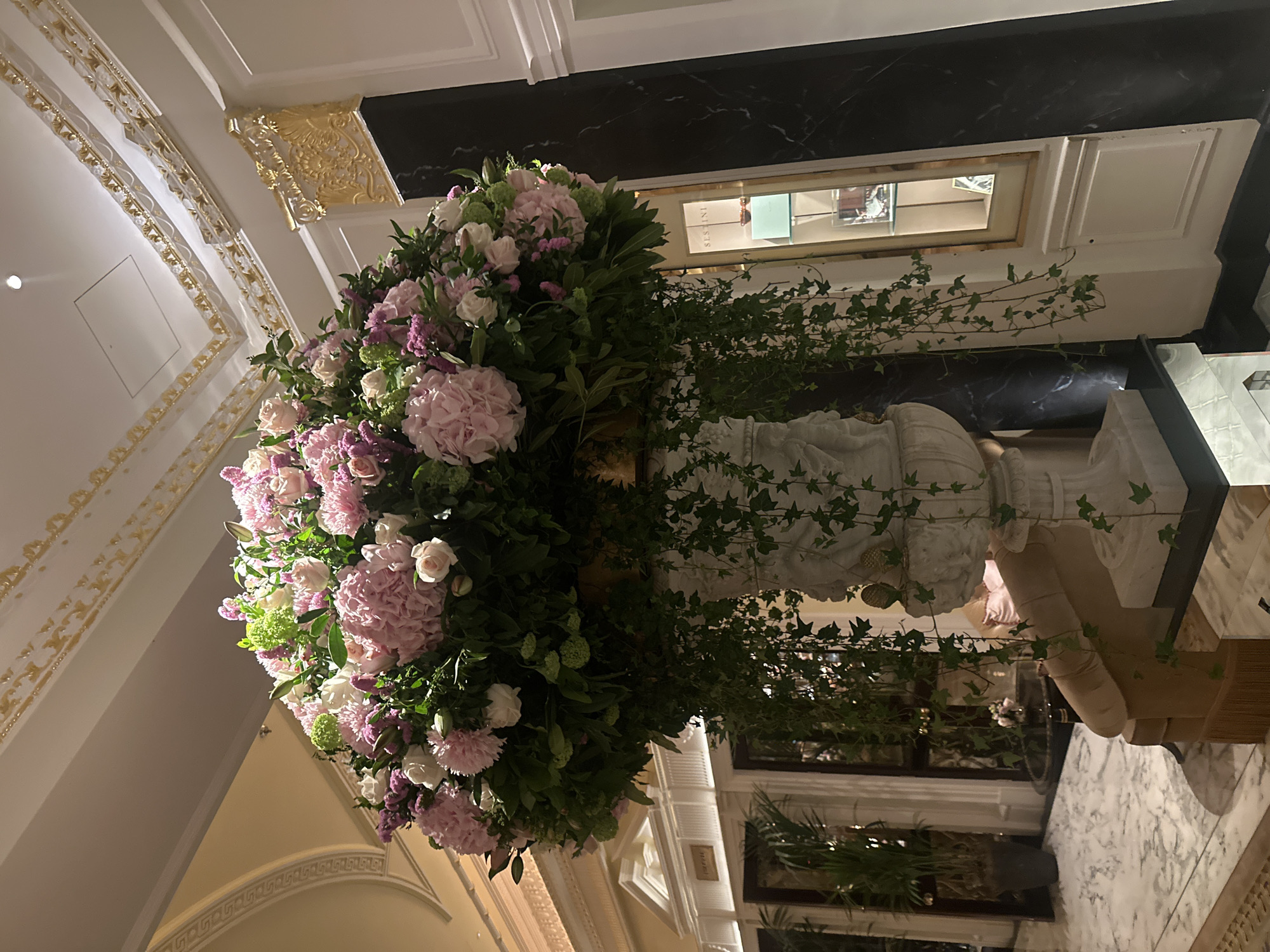 The image depicts a grand interior, likely a high-end hotel or a luxury retail space in London, United Kingdom. The main subject is a large, ornate floral arrangement consisting of pink and white hydrangeas, roses, greenery, and trailing ivy. The flowers are arranged in a classical-style urn or vase, possibly made of marble or a similar material. The vase rests on a table.
The background features architectural details characteristic of London, including decorative cornices, moldings, and arched doorways. The color palette is dominated by white, gold, and marble, suggesting a luxurious and traditional setting. A dark marble element with gold accents sits above the flower arrangement. The overall impression is one of elegance, grandeur, and sophistication.