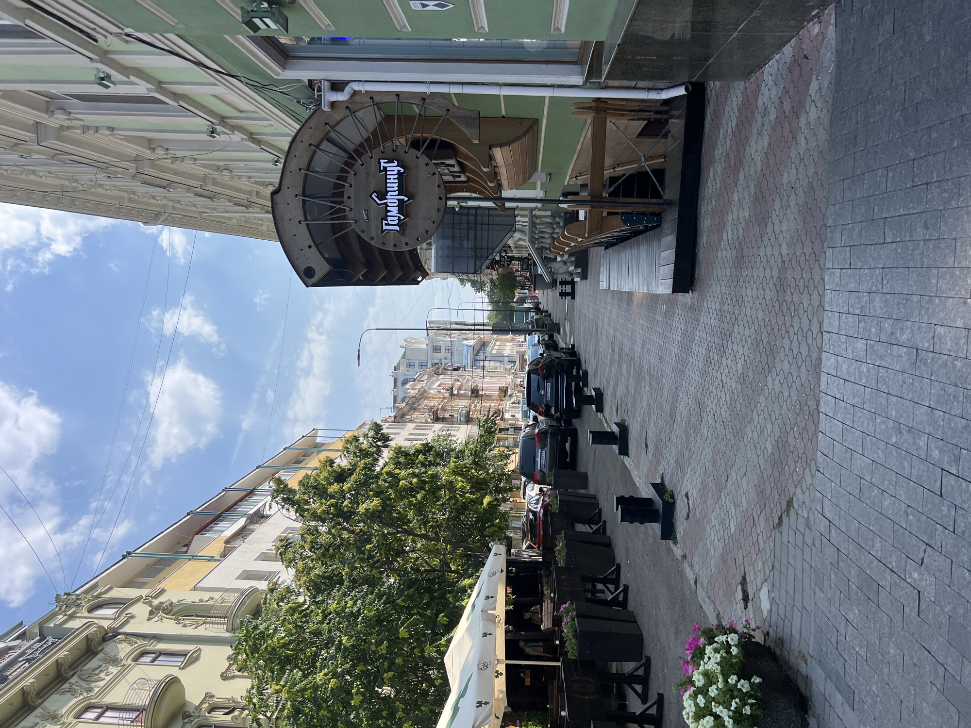 The image depicts a street scene in Odesa, Ukraine, on a bright, partly cloudy day. The perspective is from street level, looking down a paved sidewalk and street lined with buildings and trees. On the left side of the street, there is an outdoor seating area for a restaurant or cafe, covered by a white umbrella with branding. Black planters with flowers are arranged along the sidewalk in front of the seating. Several cars are parked along the street.
The buildings on both sides of the street exhibit architectural details characteristic of Odesa. The building on the left has a light yellow facade with ornate balconies and decorative elements. The building on the right is painted green and features a distinctive circular wooden sign with Cyrillic lettering that reads "Гамбринус" (Gambrinus). The street pavement is a patterned brickwork in shades of red and grey.
In the background, more buildings are visible, some with balconies and intricate facades, suggesting a historic urban environment. Power lines are visible crisscrossing the sky. No people are clearly identifiable in the image. The scene is static, capturing a typical urban streetscape.