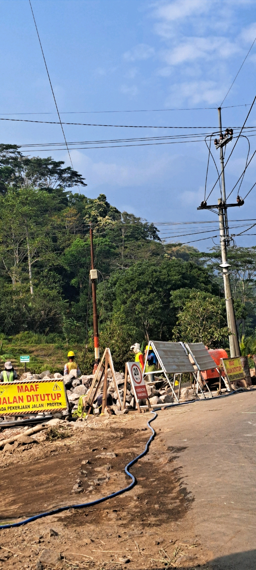 In Duyung, Indonesia, a road construction site is depicted with a section of the road closed off. A bright yellow sign with black text, reading "MAAF JALAN DITUTUP ADA PEKERJAAN JALAN / PROYEK," indicates the closure due to road work. Several workers in safety vests and hard hats are present, engaged in the construction activities. One worker is standing near the yellow sign, another is seen in the background near some scaffolding, and a third individual in a hard hat is visible further back. In the foreground, a blue hose lies on the dirt and asphalt surface. Barriers made of wood and rocks are erected to delineate the closed area. A red piece of construction equipment, possibly a small generator or pump, is situated behind the barriers. The scene is set against a backdrop of lush green trees and a bright blue sky with scattered clouds. Overhead, power lines are visible, suspended from utility poles.